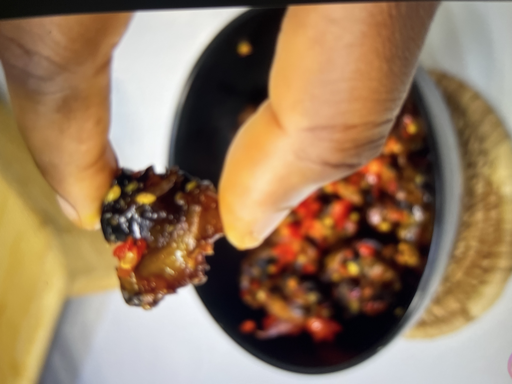 A close-up shot shows a hand holding a piece of food over a bowl filled with similar food items. The food is dark brown, glistening, and appears to be mixed with small red and yellow pieces, likely chilies and possibly corn. The hand is light-skinned, and the fingernails are visible. The bowl is black and rests on a woven placemat. The background is blurred, with a wooden surface visible at the top and a white surface below. The scene is brightly lit, suggesting an indoor setting. There is no specific information or visual cues in the image to indicate a location in Abuja, Nigeria.