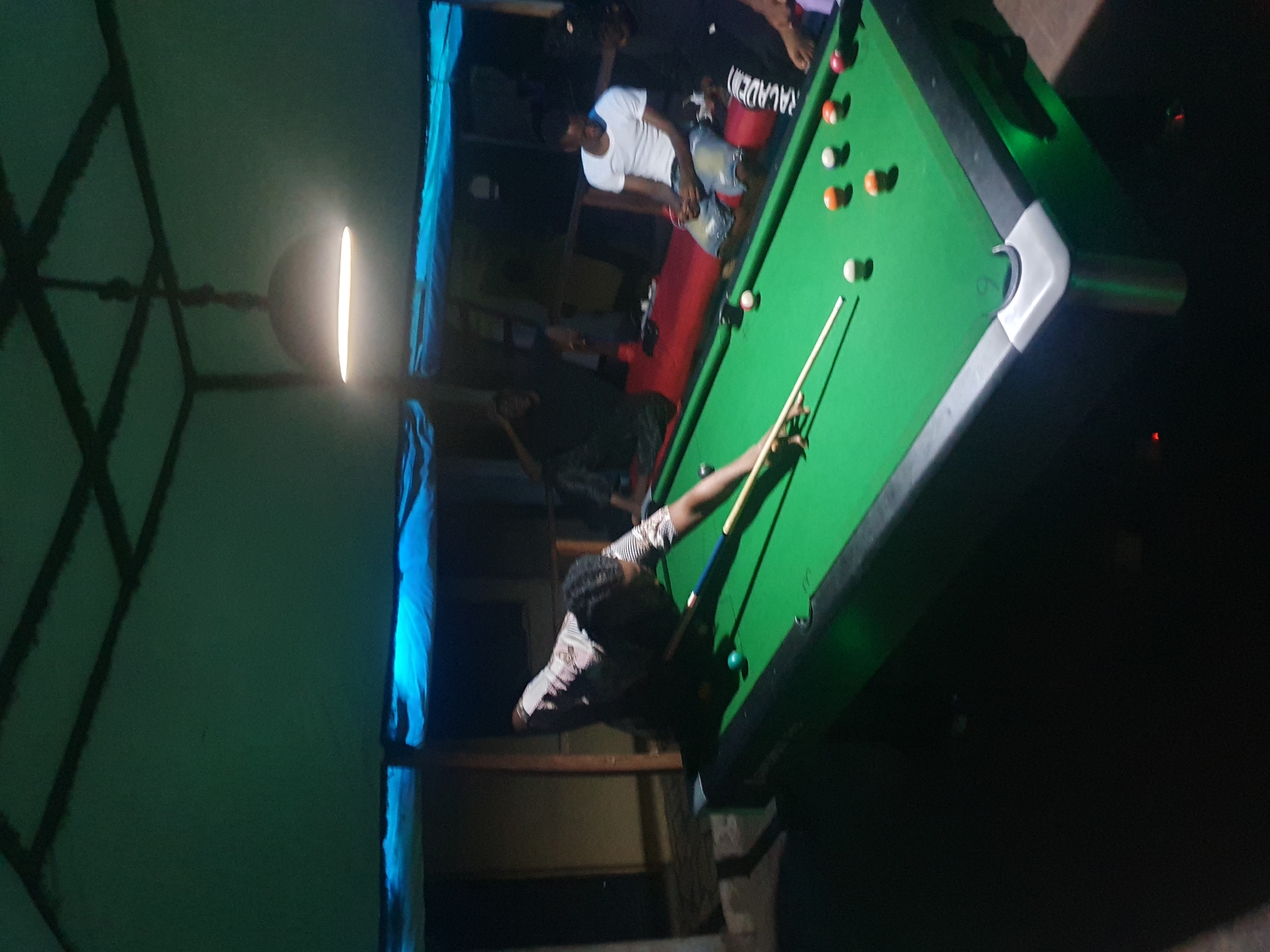 This image captures a nighttime or evening scene of people playing pool (billiards) in what appears to be a casual indoor setting, possibly a game room or bar in Lokoja, Nigeria.
The primary subject is a man in the foreground, positioned to take a shot at a bright green-felt pool table. He is holding a pool cue, aiming at the balls scattered across the table. Several pool balls, including solid and striped ones, are visible on the table's surface. Another pool cue rests on the table near the balls.
Around the table, other men are present, some standing and observing the game. One individual wearing a white t-shirt and light-colored shorts is seen sitting on what looks like a red bench or cushion on the far side of the table. Another person's legs are visible at the top left. The general atmosphere appears relaxed and social.
A dominant feature of the scene is the lighting. A circular light fixture with a prominent glowing strip hangs directly above the pool table, illuminating it brightly and casting a strong glow. The rest of the room is dimly lit, enhancing the focus on the game. To the far left, a blue-lit area or fabric adds a cool tone to the background, and dark, angular structural elements (possibly part of a ceiling or tent) cast shadows on the wall.
Visible text on the back of one person's attire (shorts or trousers) near the top of the pool table appears to read "RALADEN" or a similar partial word. The image is rotated, presenting a somewhat diagonal perspective of the scene.