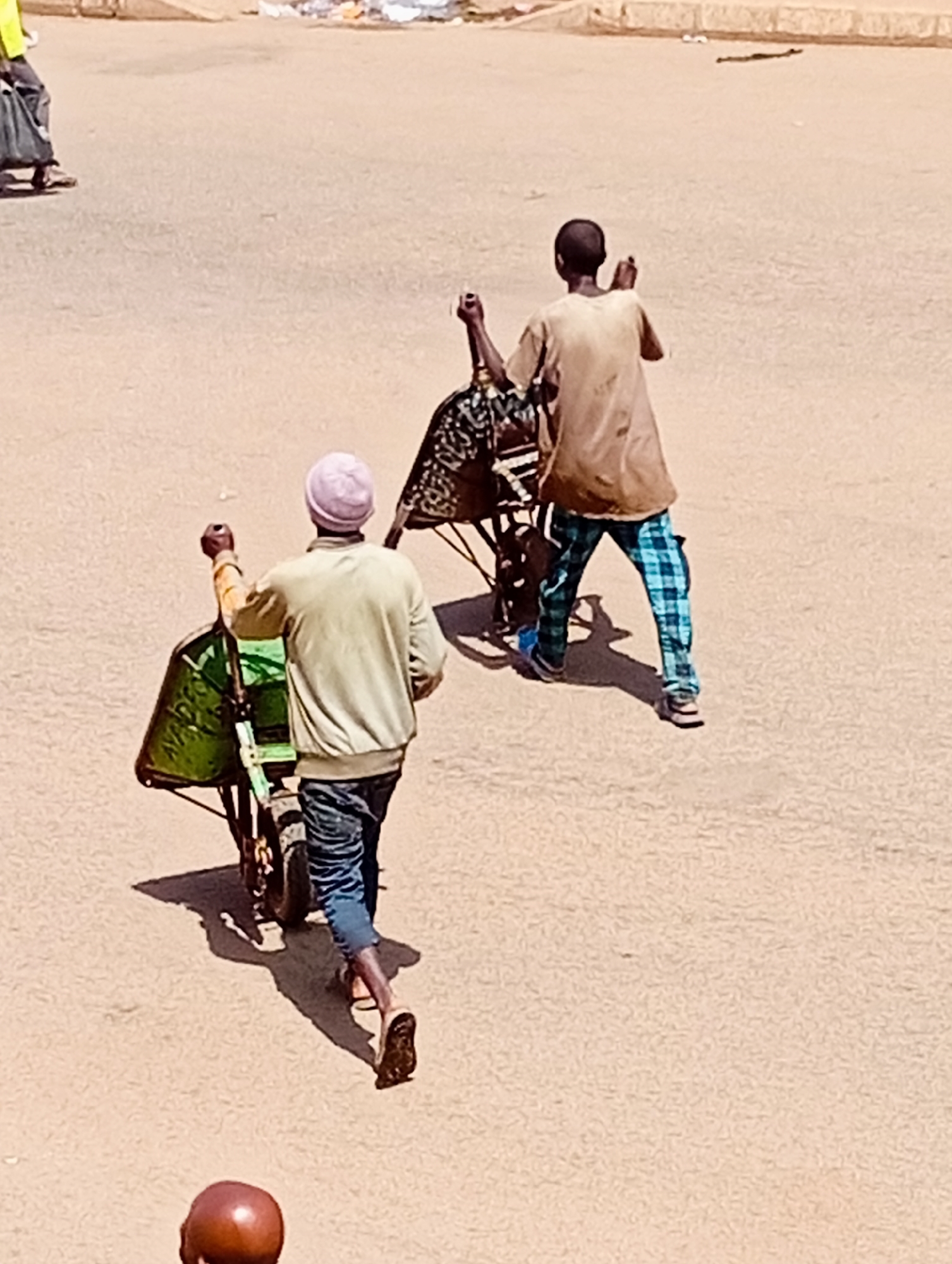 In Bukuru, Nigeria, two individuals are seen in a dusty, outdoor environment, likely a street or open market. One person, wearing a light-colored jacket, a pink head covering, and blue jeans, pushes a green wheelbarrow filled with a black tarp. The wheelbarrow has two wheels and a single handle. This individual is walking away from the viewer, with their right arm raised.
Behind them, another person, dressed in a tan t-shirt and blue and green checkered pants, is also walking away. They are pushing a wheelbarrow containing another black tarp, and their arms are raised. The ground appears to be a dirt or unpaved surface, with shadows cast by the people and the wheelbarrows. In the background, there are faint signs of activity and possibly some debris. A reddish-brown spherical object, possibly a head, is visible in the lower foreground.