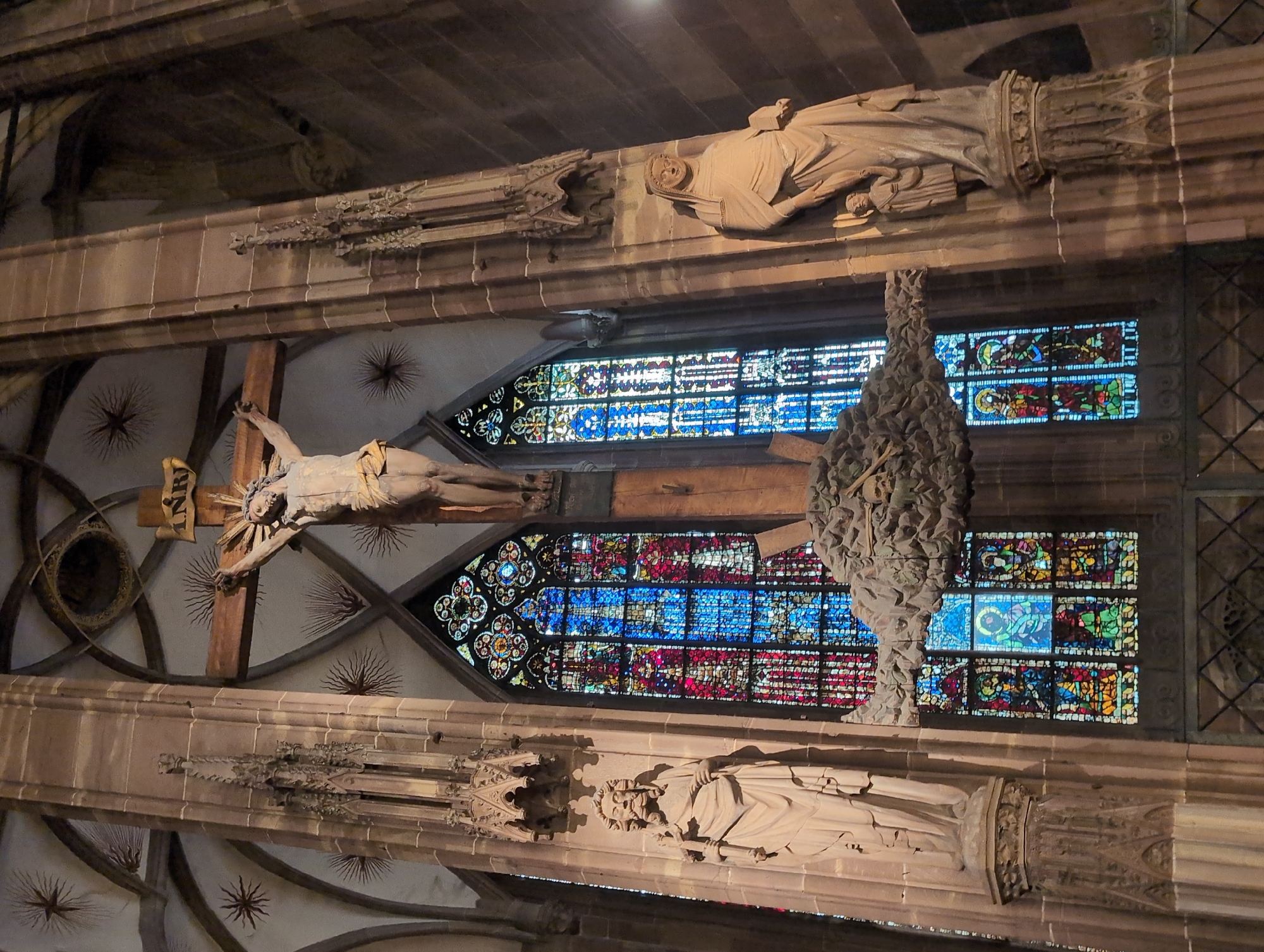 The image depicts the interior of the Reims Cathedral in Reims, France. A large wooden crucifix bearing the figure of Jesus Christ is suspended centrally. The crucifix is adorned with a halo-like sunburst behind Jesus' head and a sign labeled "INRI". To the left and right of the crucifix stand two large stone statues on pedestals. The statue on the left depicts a bearded male figure holding a staff, and the statue on the right shows a robed female figure holding a smaller figure. Behind the statues and crucifix, large, ornate stained-glass windows fill the space with vibrant colors, primarily blues, reds, and yellows. The architectural style is Gothic, evident in the pointed arches of the ceiling and the decorative stonework. The overall atmosphere is solemn and grand, typical of a cathedral interior.