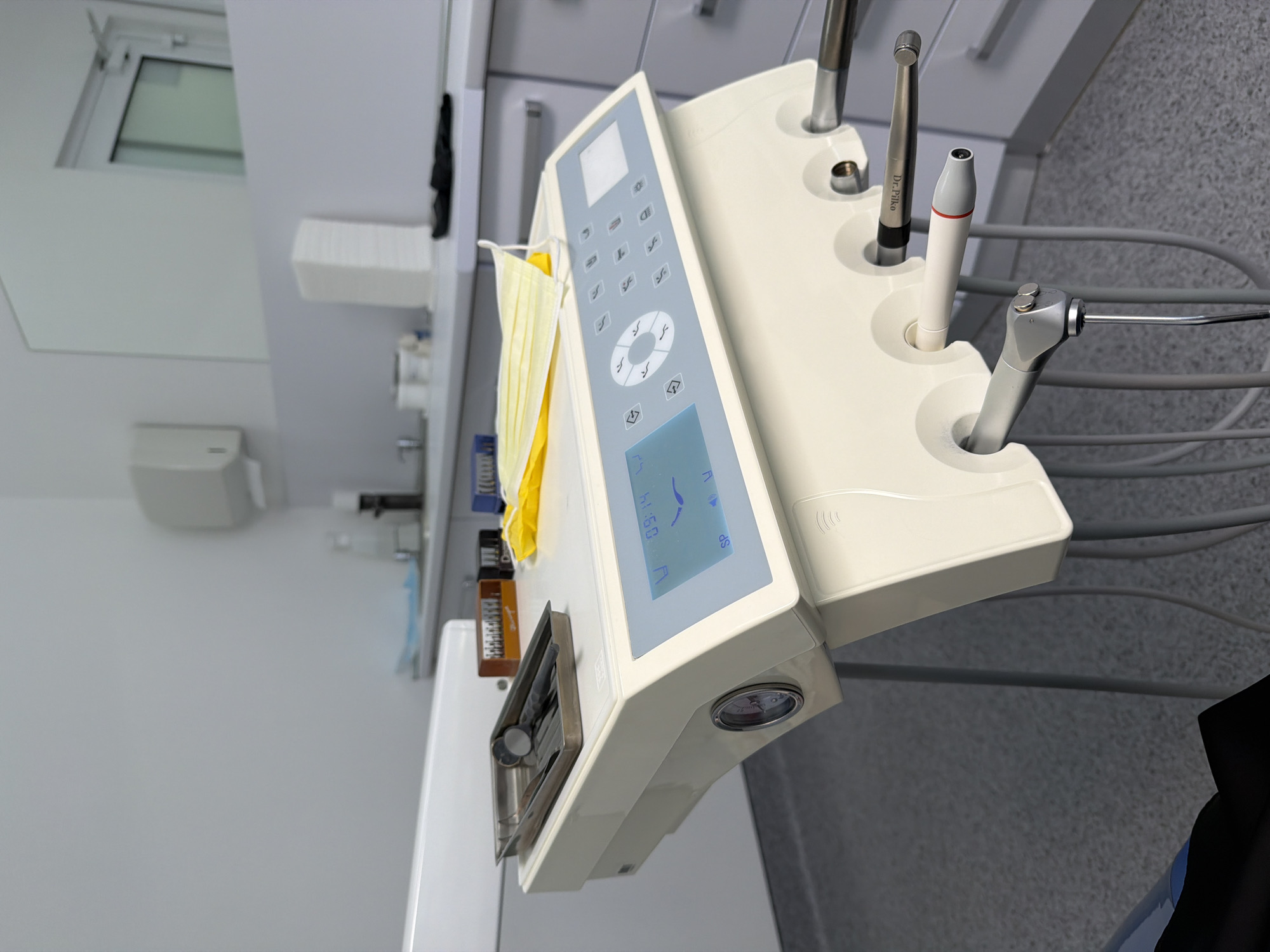 In an indoor setting, possibly a dental clinic, a modern, off-white dental unit is positioned in the foreground. The unit features a digital display screen showing the time "09:14" and various icons, along with a circular control panel. Below the control panel, there are several ports where dental handpieces are stored, with three visibly inserted: a high-speed handpiece with a silver and grey metallic finish, a white handpiece with a red stripe, and another metallic handpiece labeled "Dr. Piko."
On the top surface of the dental unit, a metal tray holds dental instruments, including what appear to be a mirror, explorer, and probe. A folded yellow surgical mask rests next to the instrument tray.
The background reveals a clean, minimalist environment with white walls and grey cabinetry. A sink with a faucet is visible in the upper left, and a dispenser is mounted on the wall. A large mirror reflects a window and a partially open door in the upper right portion of the image. The floor is covered in a speckled grey material. The scene appears to be a well-equipped and tidy dental operatory.