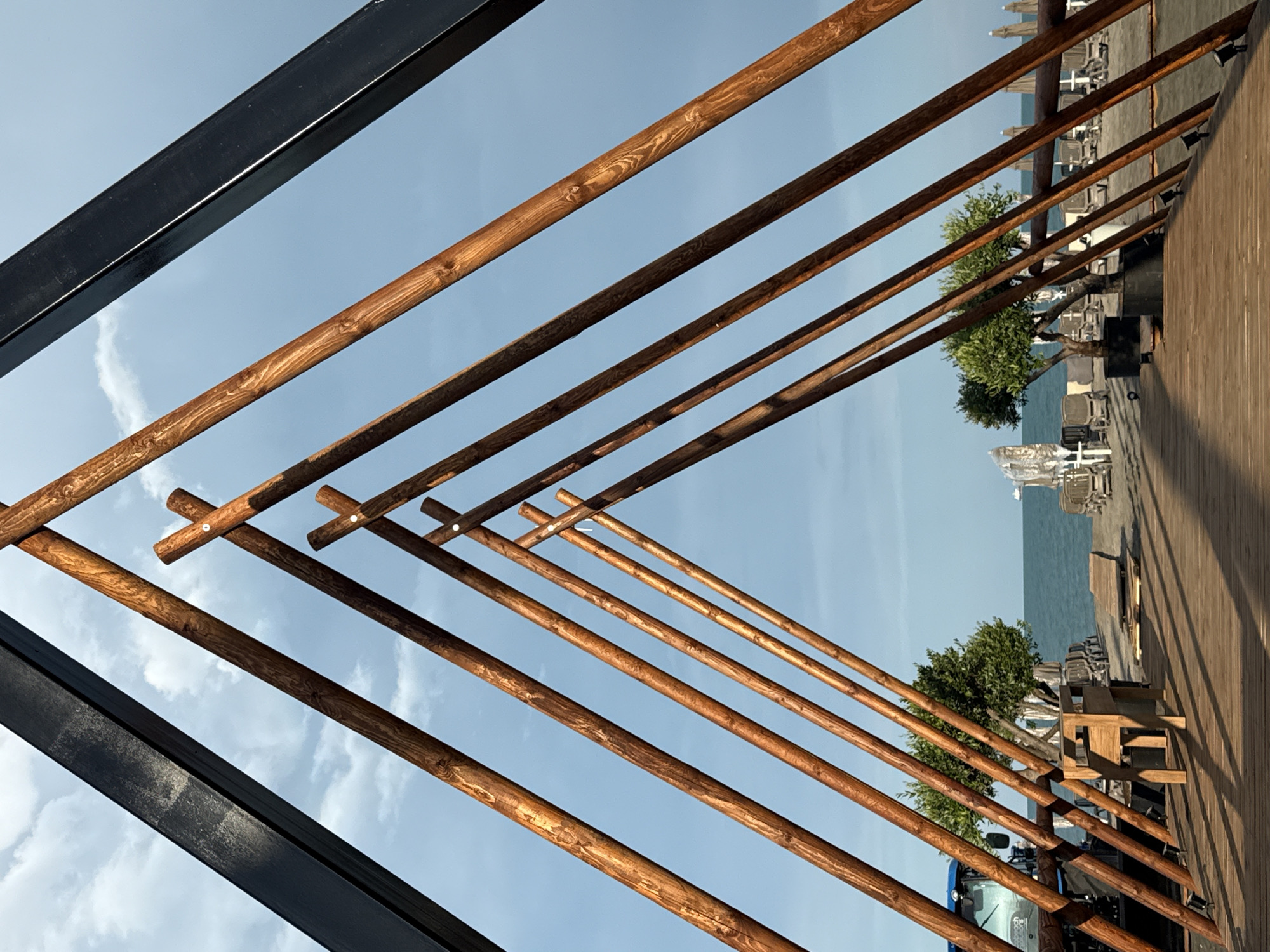 The image captures a view looking up at a wooden pergola structure against a bright blue sky with scattered clouds. The pergola is constructed with numerous wooden beams, some intersecting at angles to create a triangular pattern. A thick, black metal beam is visible in the upper left corner, likely part of the main support for the structure.
Beyond the pergola, a wooden deck is visible, with rows of empty lounge chairs and beach umbrellas suggesting a daytime scene at a resort or beach club. The sea is visible in the background, with a distant tree and some outdoor furniture. A blue tractor is partially visible in the lower left, adding a touch of industrial or operational context to the scene. The lighting indicates it is daytime, possibly late afternoon, with shadows cast by the pergola beams. The overall impression is one of a well-maintained outdoor leisure space, possibly in Burgas, Bulgaria, as indicated by the location context. There is no visible text in the image.