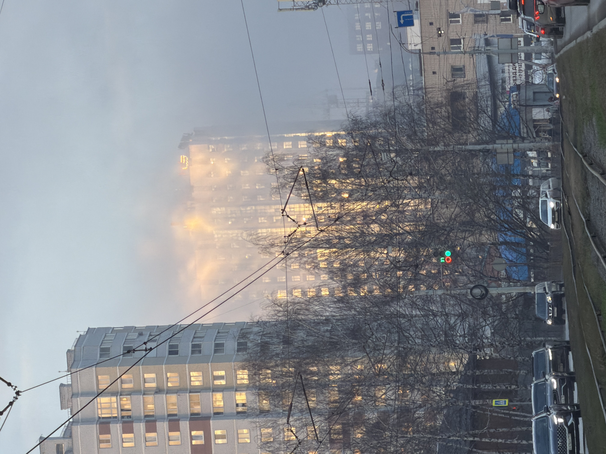 The image shows a street scene in Izhevsk, Russia, with buildings visible through a hazy or foggy atmosphere. The buildings are tall, with rows of windows reflecting sunlight. Bare trees obscure the view in the foreground. Overhead tram lines are strung across the sky. The right side of the image shows a road with cars and a grassy verge. A traffic light is visible with a green signal illuminated. Pedestrian crossings are indicated.