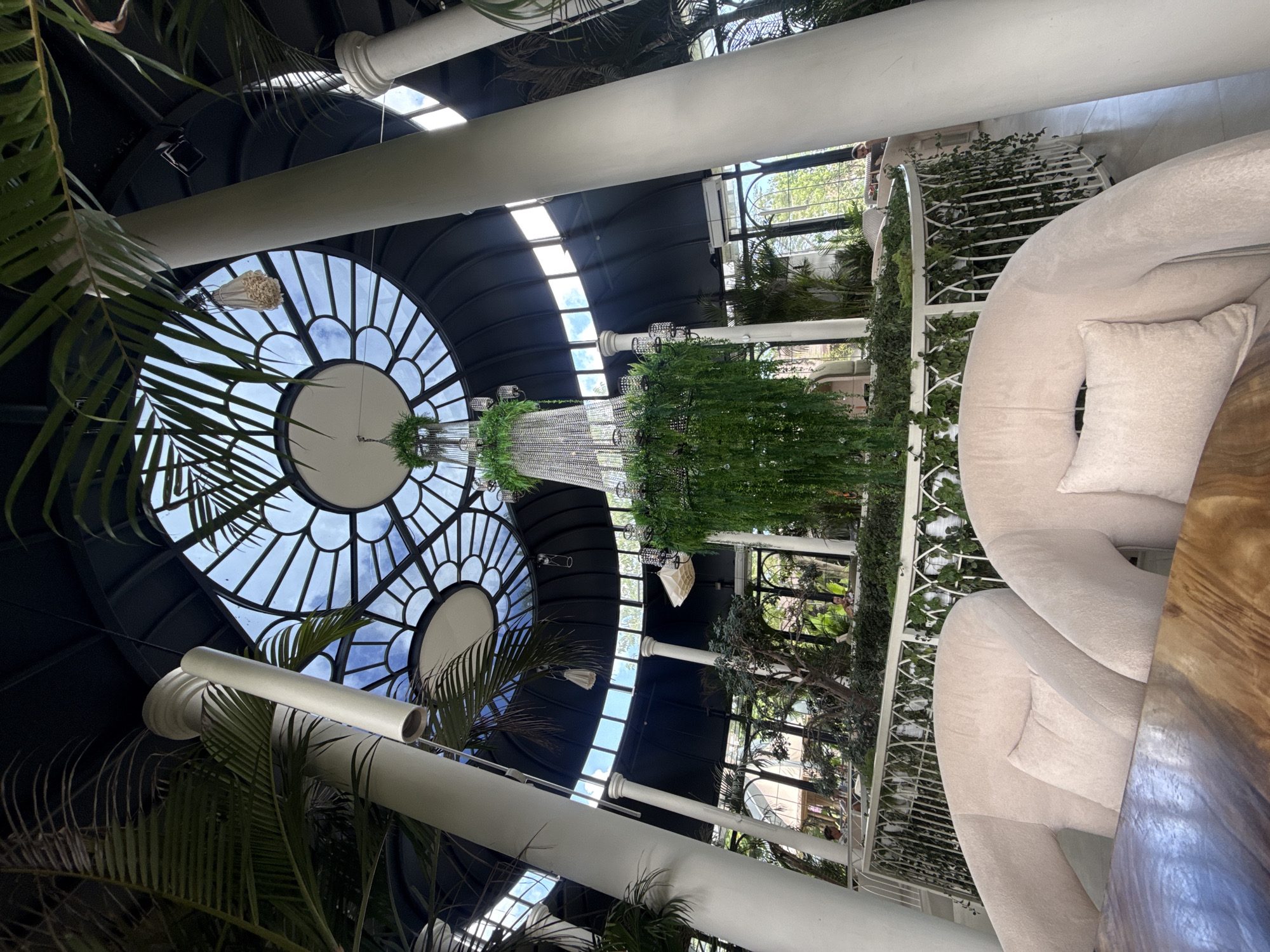The image shows an interior view of a building, likely in North Kuta, Indonesia, given the prevalence of tropical plants. The architecture is characterized by a large, circular glass ceiling with a radial pattern, allowing natural light to flood the space. Tall, white pillars support the structure, adding to the grandeur. Lush, green plants are abundant, both around the pillars and hanging from a chandelier-like structure in the center of the room. A white, curved sofa with a pillow is visible on the right side of the image, offering seating within the space. The scene suggests a luxurious or upscale setting, possibly a hotel lobby or restaurant. No individuals are clearly visible.
