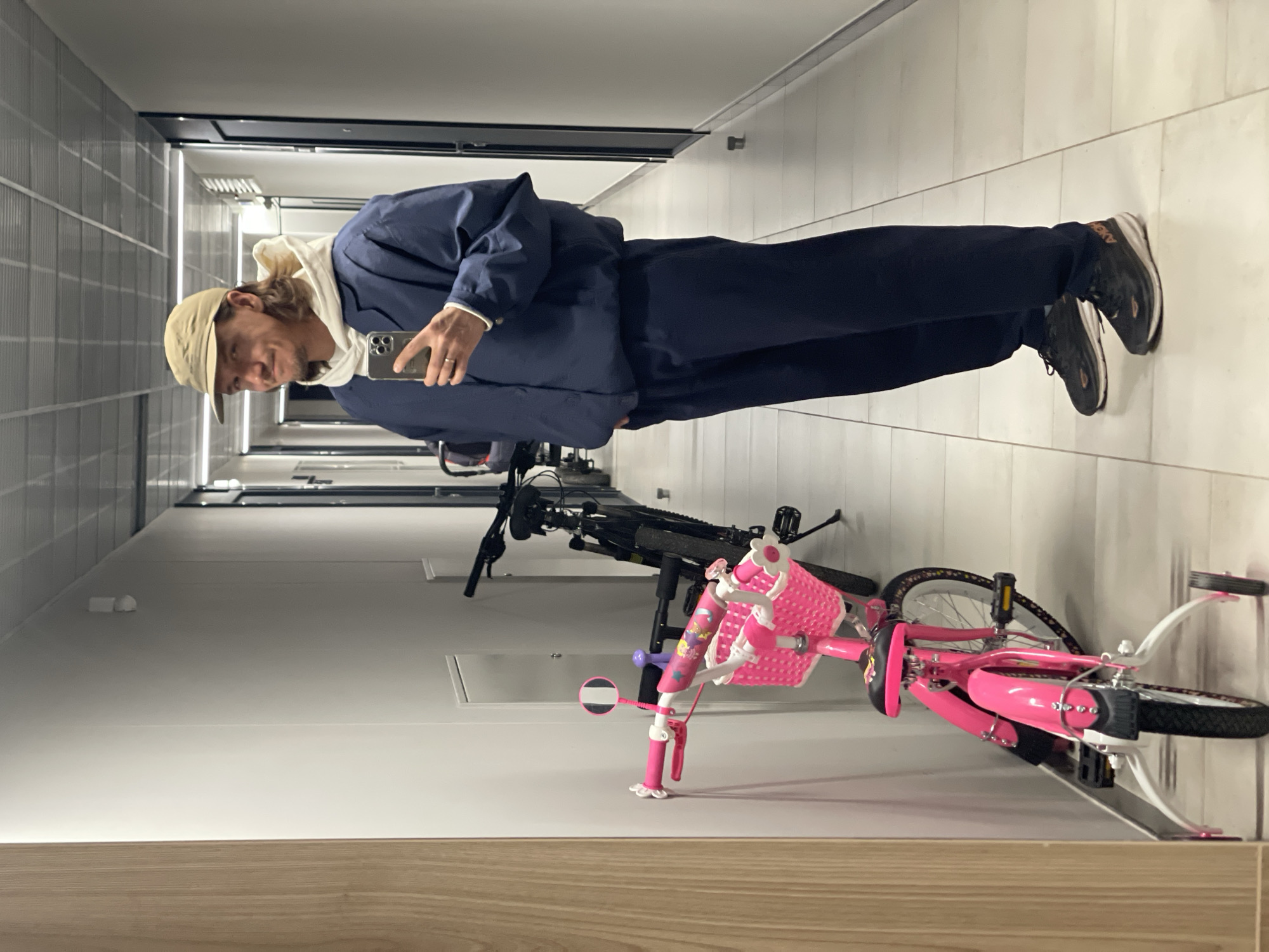 A person wearing a blue jacket, dark pants, sneakers, and a beige cap is taking a selfie in a hallway. The hallway has a white tiled floor and white walls. There are two bicycles in the hallway, a pink child's bicycle and a black mountain bike. The location is unknown.