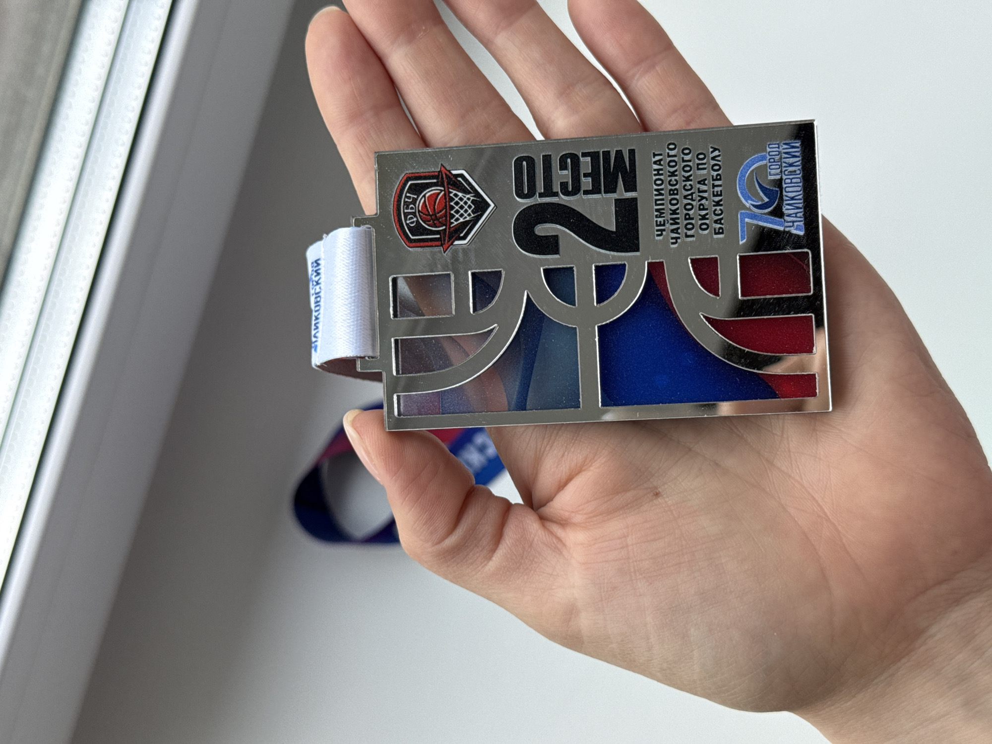A person's right hand holds a rectangular, silver-colored metal medal. The medal commemorates second place ("MECTO 2") in the Chaikovsky urban district basketball championship. It also references the 70th anniversary of the city of Chaikovsky, Russia. The medal features a basketball net logo on the left. The medal hangs from a white and blue ribbon with lettering in Russian. The hand and medal are positioned near a white window frame.