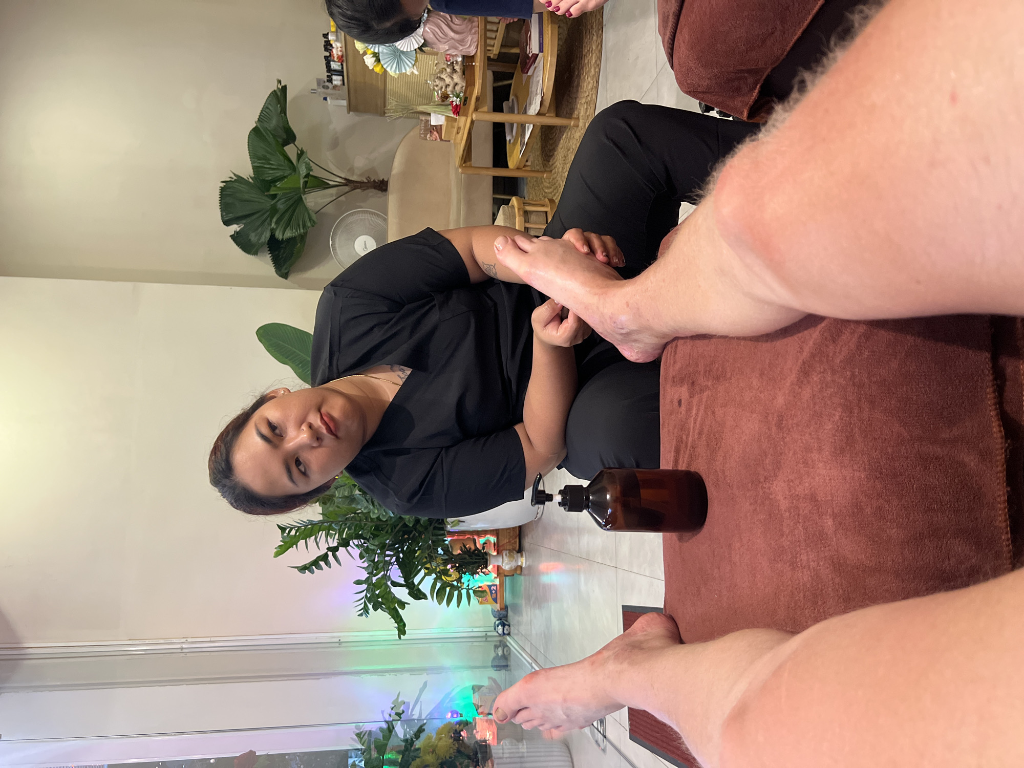 A woman in Bac Nha Trang Ward, Vietnam, dressed in a black top and pants, is providing a foot massage to a person lying on a brown upholstered massage table. A bottle of massage oil rests near the person's feet. A window with plants in front of it is visible in the lower left corner of the image.