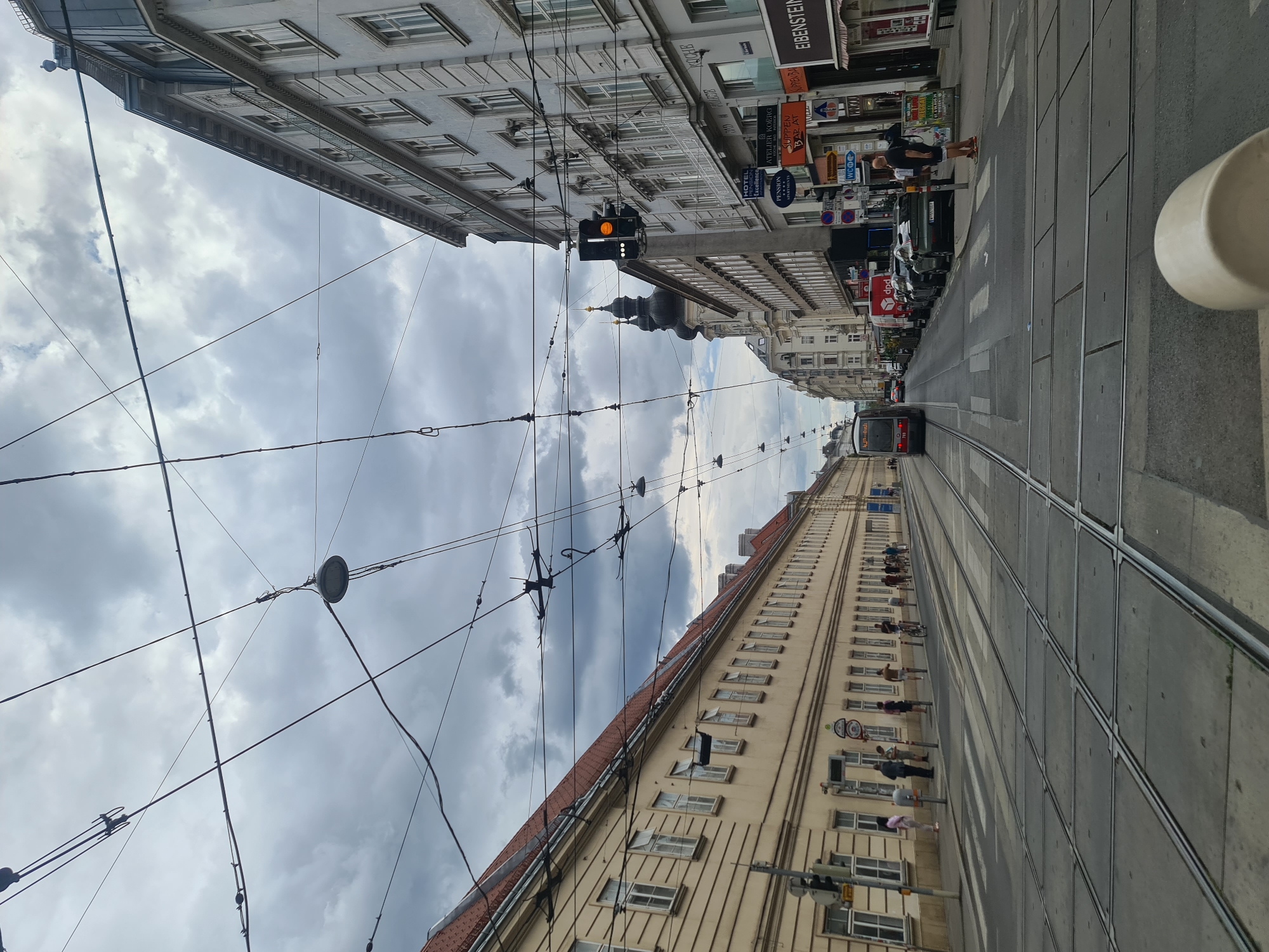 The image depicts a street scene in Vienna, Austria, likely near the city center, characterized by the presence of tram tracks and overhead electrical wires. The perspective is from a street level, possibly from a moving tram or a location close to the tracks.
On the left side of the street, a long, light yellow building with numerous windows stretches into the distance. The building has a red-tiled roof and appears to be residential or commercial. Several individuals are seen walking or standing on the sidewalk in front of this building.
On the right side of the street, taller, multi-story buildings line the sidewalk. These buildings are a light gray or white color and have architectural detailing, including balconies and rows of windows. A prominent traffic light, showing an orange/yellow light, is visible hanging over the street. Shops and businesses are indicated by signs, with one sign reading "EIBENSTEIN" and another for "HOTEL PENSION EXCELLENCE." Several vehicles are parked along the right side of the street, including cars and a delivery van with "dpd" branding.
A red tram is visible in the middle distance, moving away from the viewer. Tram tracks are laid out prominently in the foreground and extend down the street, indicating an active tram line. The sky is overcast with dark clouds, suggesting a cloudy or potentially rainy day. The overall atmosphere is urban and bustling, with a mix of architectural styles and daily urban activities.