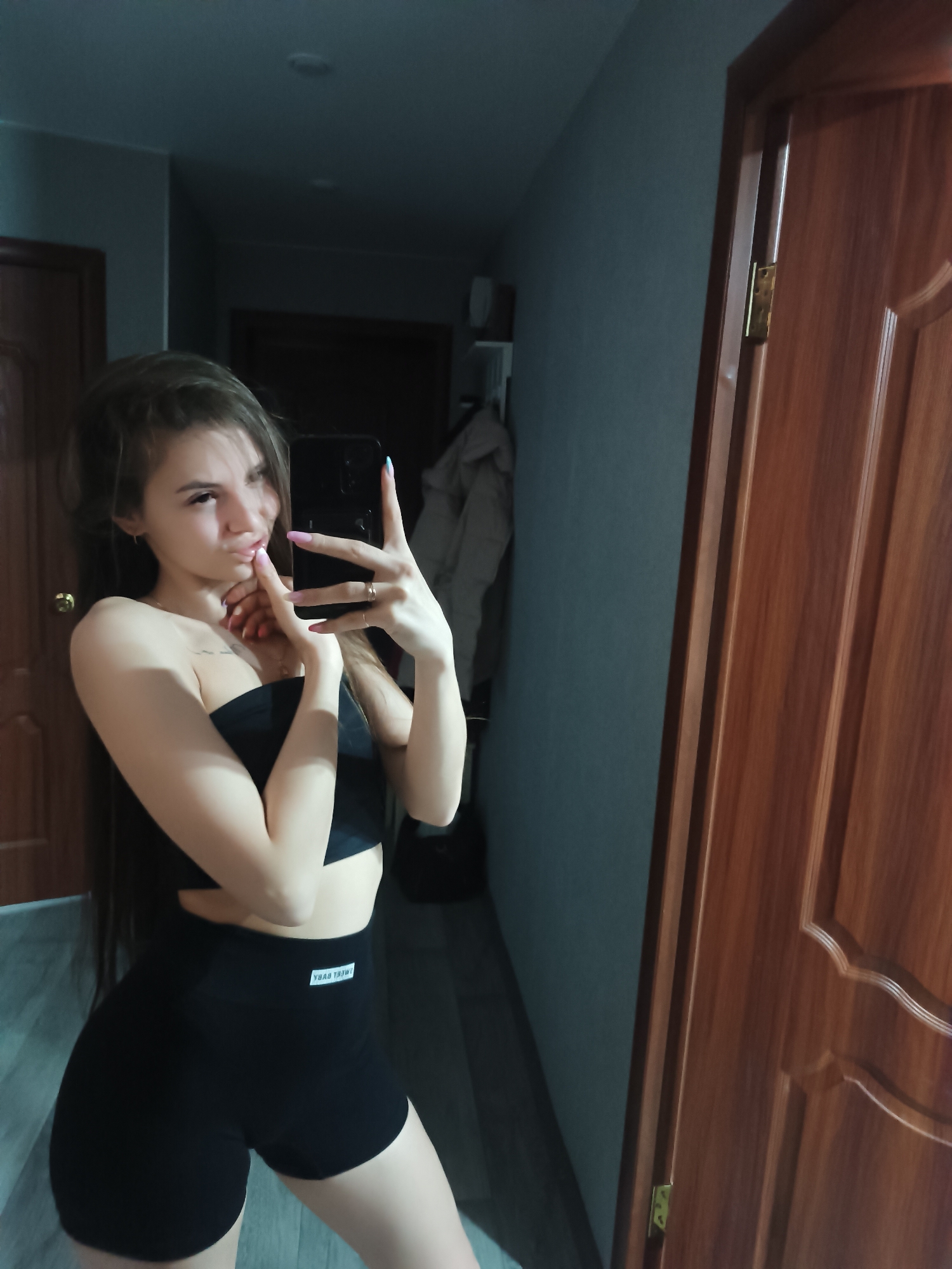 A young woman is taking a selfie in an indoor setting, likely a hallway. She is wearing a black strapless top and black shorts. The background includes a hallway with doors and a coat hanging. It is possible that the location is Chaikovsky, Russia, but this cannot be confirmed from the image alone.