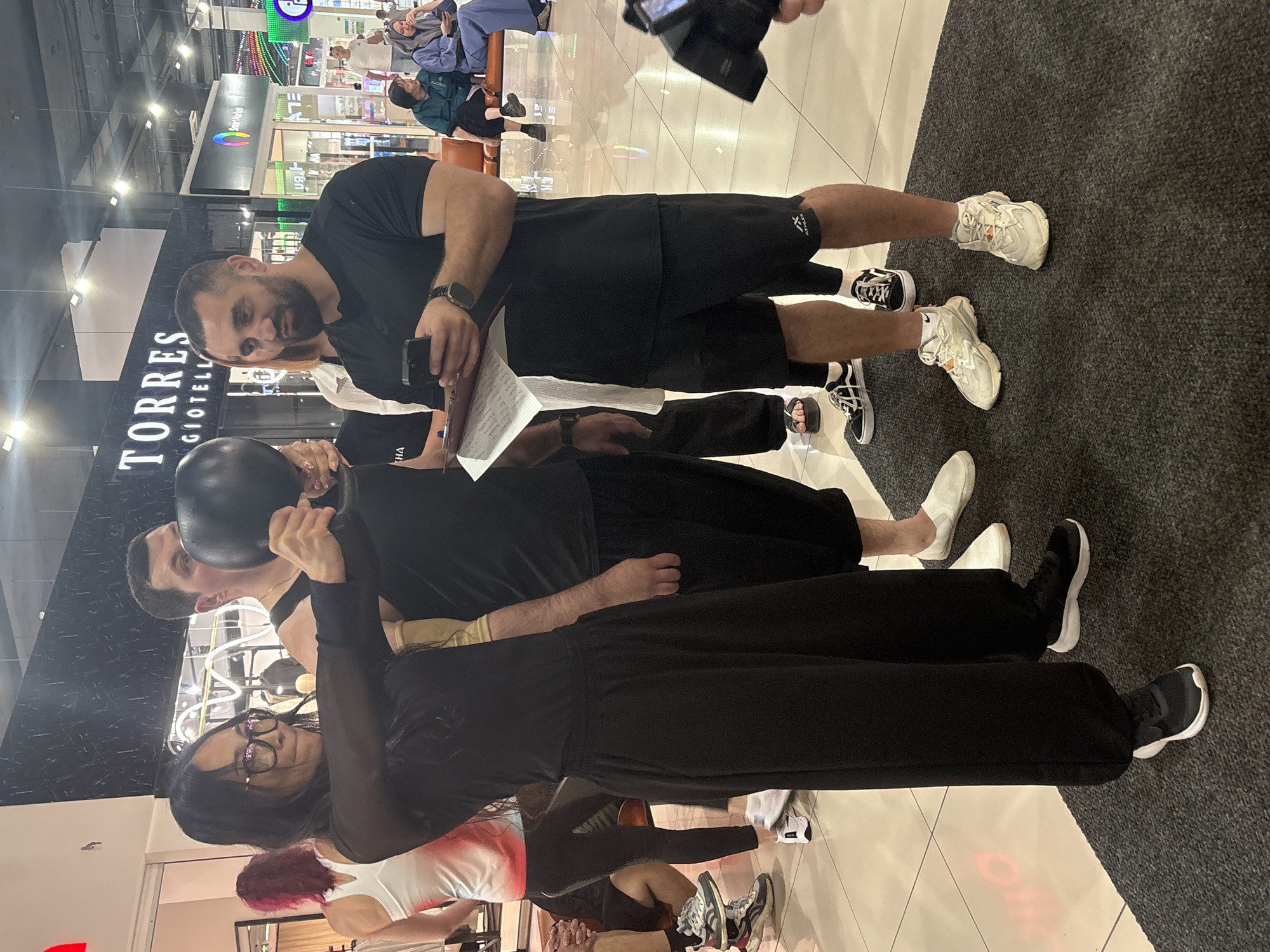 The image shows a group of people indoors, possibly in a shopping mall. In the foreground, a woman with long dark hair and glasses is holding a black kettlebell. She is wearing a black sheer long-sleeved top and black loose-fitting pants. To her right, a man with dark hair and a beard, wearing a black t-shirt and black shorts, is looking at his phone and holding a clipboard. Behind them, other individuals are visible. The floor is tiled and reflective. A large sign that reads "TORRES GIOIELLI" is visible in the background. There are also displays of merchandise and people sitting on benches in the distance. A camera is visible in the lower right corner of the image.