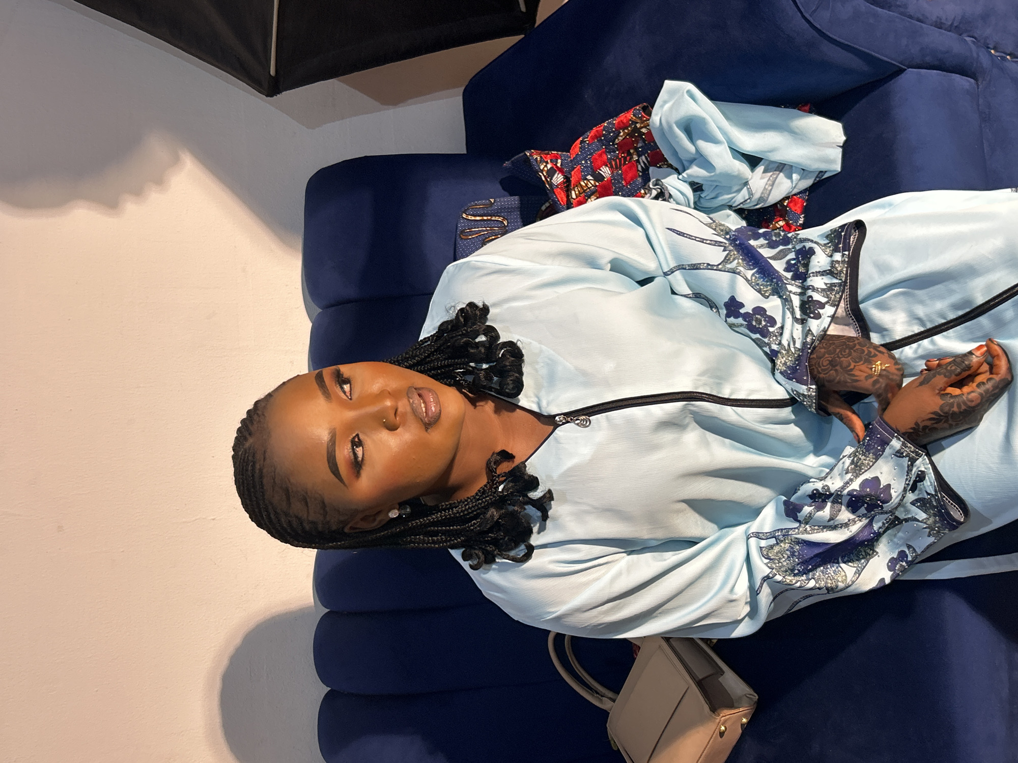 A young woman with braided hair and henna tattoos on her hands is lying on a blue velvet couch. She is wearing a light blue dress with floral patterns on the sleeves. There is a handbag next to her. The setting appears to be indoors, with a white wall partially visible in the background. There is no verifiable information in the image to indicate it was taken in the city of Jos, Nigeria.