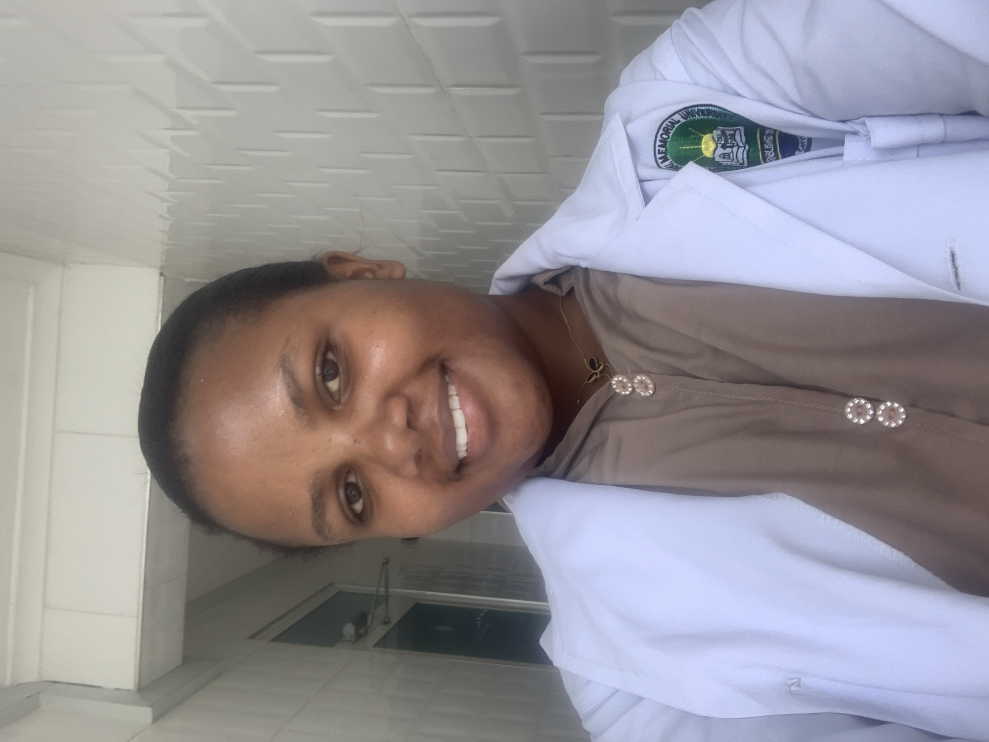 The image displays a head-and-shoulders portrait of an adult female located in what appears to be a brightly lit indoor or semi-indoor corridor at Kairuki Memorial University in Dar es-Salaam, Tanzania. The subject has dark skin, dark hair pulled back, and is smiling directly at the camera, revealing white teeth. She is wearing a white lab coat, which has the emblem of "Kairuki Memorial University" visible on the right lapel. Underneath the lab coat, she wears a light brown or taupe collared shirt with multiple circular, decorative buttons. A gold-colored necklace with a butterfly-shaped pendant is visible around her neck. The background on the left shows a white paneled ceiling structure and a light-colored wall, with a partial view of an entryway or window in the distance. The background on the right features a white wall with a repeating raised rectangular pattern, suggesting textured tiles or panels.
