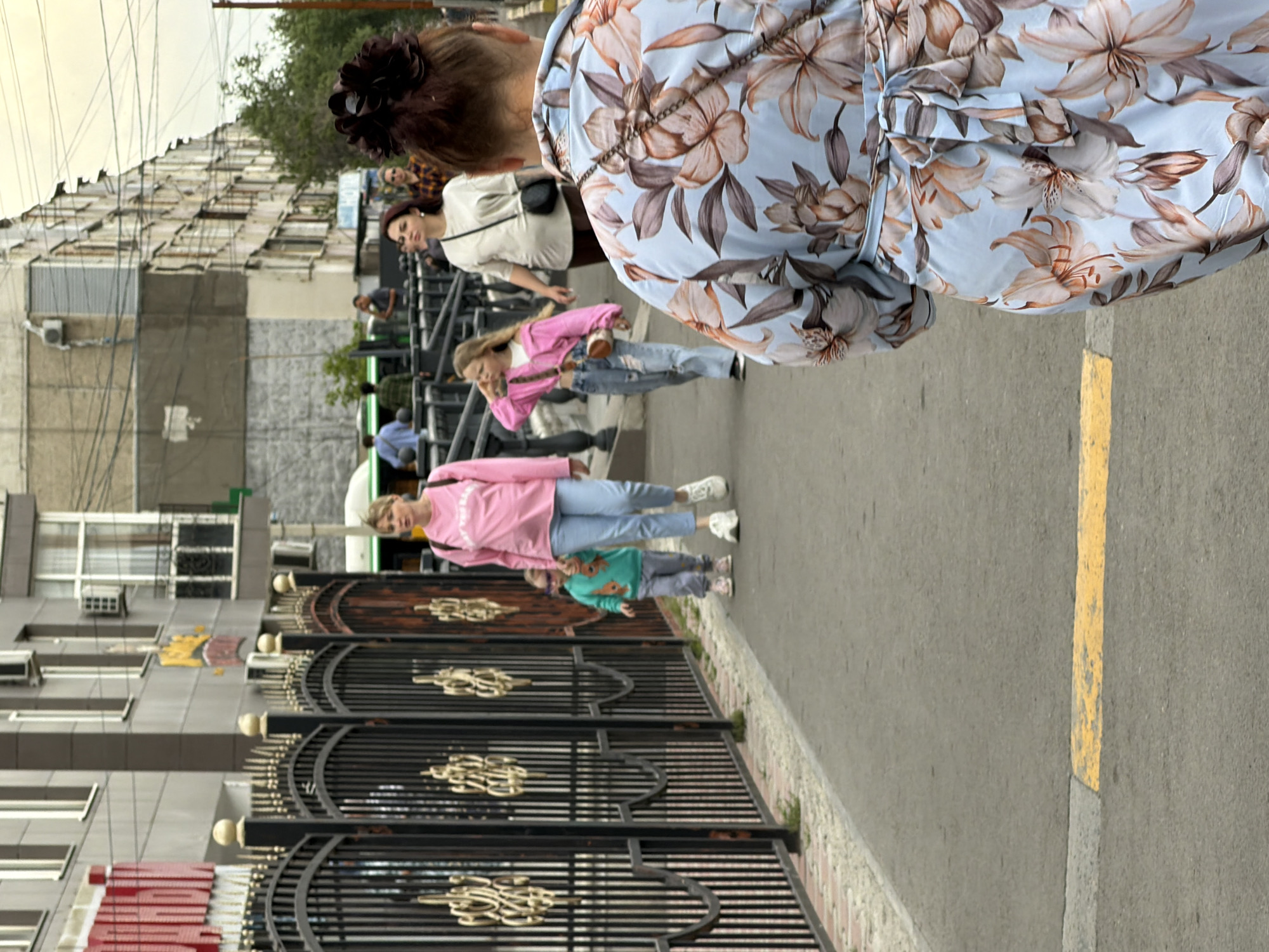 The image shows a street scene with several people walking on a sidewalk. In the foreground, a woman with her back to the camera is wearing a light blue dress with a large floral pattern. She has dark hair styled in a bun with flowers. To her left, a woman in a pink sweatshirt and light blue jeans is walking with two young children. One child is wearing a green long-sleeved shirt and gray pants, and the other is wearing a pink jacket and jeans. In the background, other people are visible walking along the sidewalk, and buildings line the street. There is a black metal fence with ornate gold details along the left side of the sidewalk. A yellow line is painted on the asphalt in the foreground.