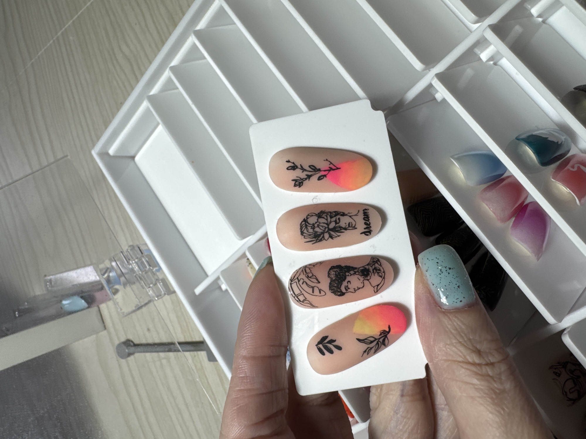 A close-up shot shows a person's hand holding a display of artificial nails. The nails are oval-shaped and feature various designs. The top nail has a black silhouette of a branch with leaves, with a gradient of pink and orange at the tip. The second nail from the top has a black outline of two faces and the word "dream" in black cursive text. The third nail features a black silhouette of two profiles facing each other, with some leafy elements. The bottom nail has a black silhouette of a branch with leaves, similar to the top nail, with a gradient of pink and orange at the tip. The artificial nails are on a white display card. In the background, a white organizer with multiple compartments holds more artificial nails in various colors and finishes, including some iridescent and translucent ones. To the left, a small clear tool with metal parts is visible on a wooden surface. The lighting suggests an indoor setting.
