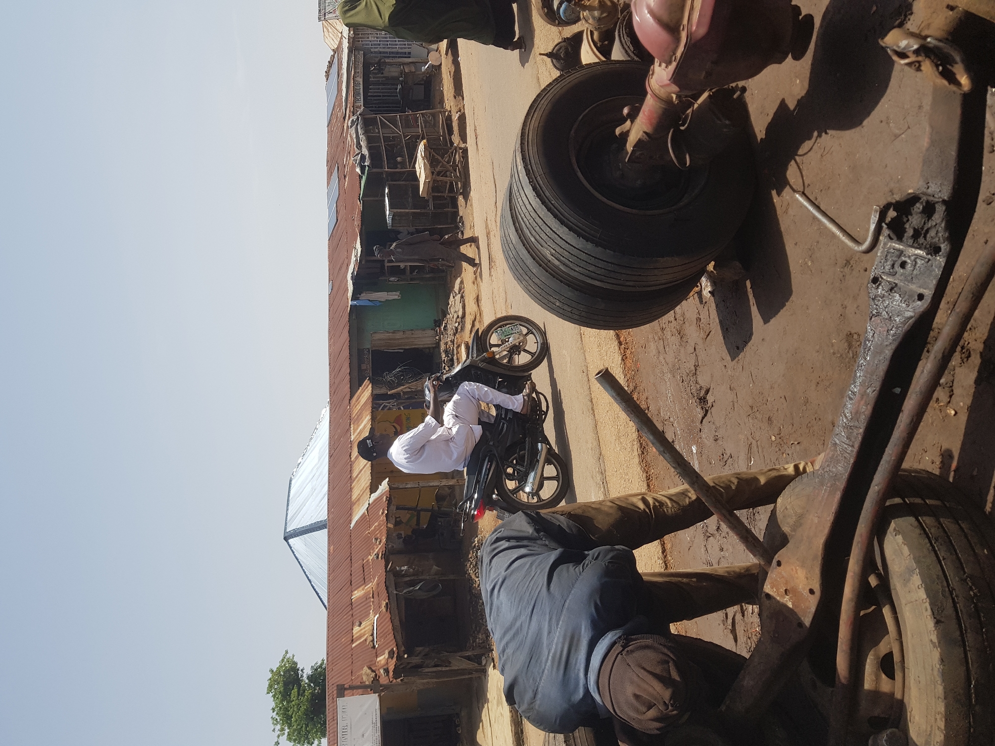 This media file depicts an outdoor scene in what appears to be a workshop or marketplace area. The primary subjects are three individuals, one of whom is riding a motorcycle. The person on the motorcycle is wearing a white traditional outfit and a black cap. Another individual, wearing a dark blue jacket and brown cap, is partially visible, hunched over what looks like mechanical parts. A third person, wearing a dark grey outfit, is standing on the left side of the frame, near a building with a corrugated metal roof.
The setting is dusty and appears to be a street with various mechanical parts, including a stack of tires and what looks like car or truck axles, scattered around. Buildings with corrugated metal roofs are visible in the background, suggesting a somewhat informal commercial area. The lighting suggests it is daytime, with sunlight casting shadows on the ground.
The activity seems to be related to vehicle repair or a vehicle parts vendor. The person on the motorcycle might be a customer or someone working in the area. The hunched-over individual appears to be engaged in some sort of mechanical work.
Notable details include the presence of a motorcycle with a license plate that reads "LSD 353 NA". The general atmosphere appears to be busy, with elements of commerce and mechanical activity. There is a visible sign on the building on the left, though the text is not entirely legible, it appears to include "TECHNOLOGY". Another sign lower down also has some text, but it is too blurry to read. The location context provided is Tukur Tukur, Nigeria.