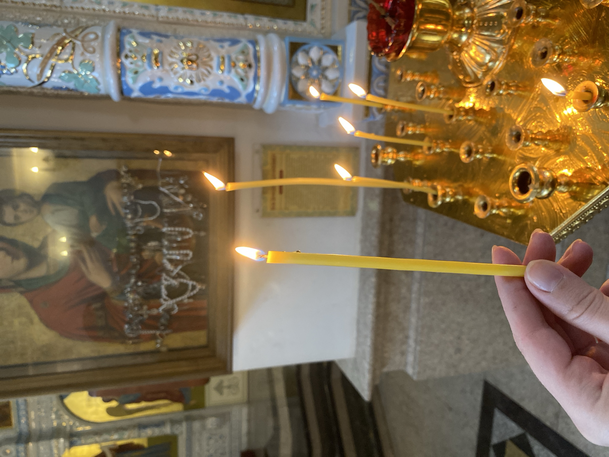 A hand holds a lit yellow candle in an Orthodox church. Multiple other candles burn in a golden candelabra. An icon of the Virgin Mary is to the left. The location may be the Church on Blood in Yekaterinburg, Russia.