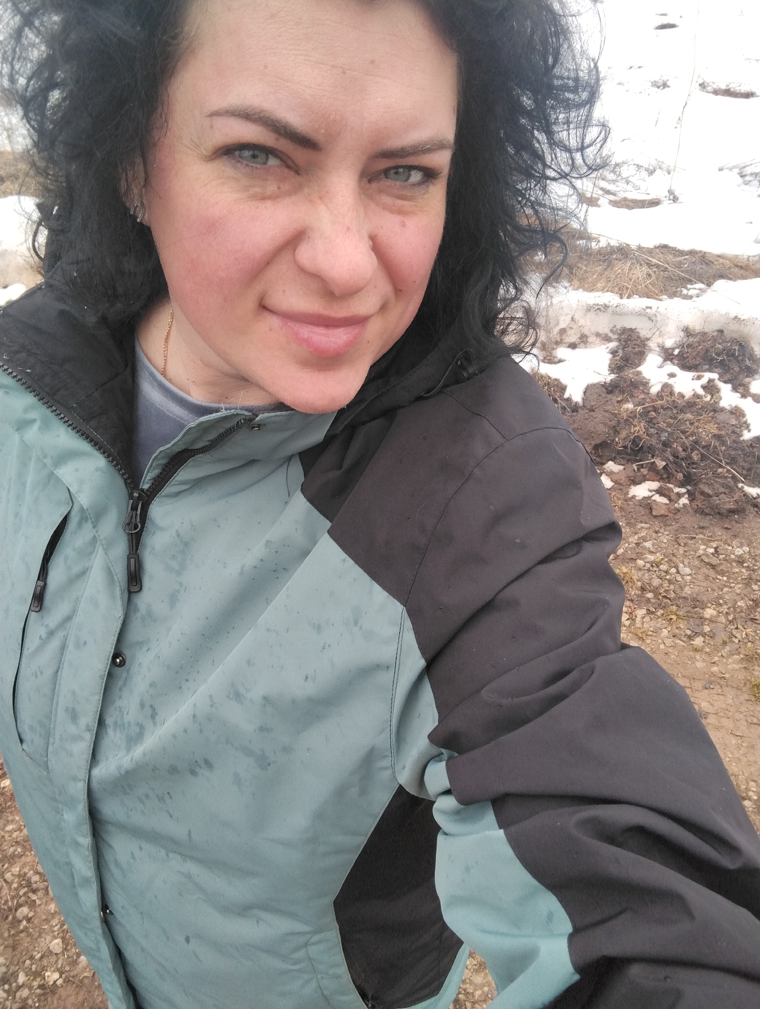 The photograph shows a woman wearing a blue-green and black jacket standing outdoors. Her dark, curly hair frames her face, and she has light-colored eyes. The jacket appears to be wet with visible water droplets. In the background, there is melting snow and muddy ground. The setting is likely a rural area based on the visible terrain and melting snow. The location of the image is Grishanki, Russia.