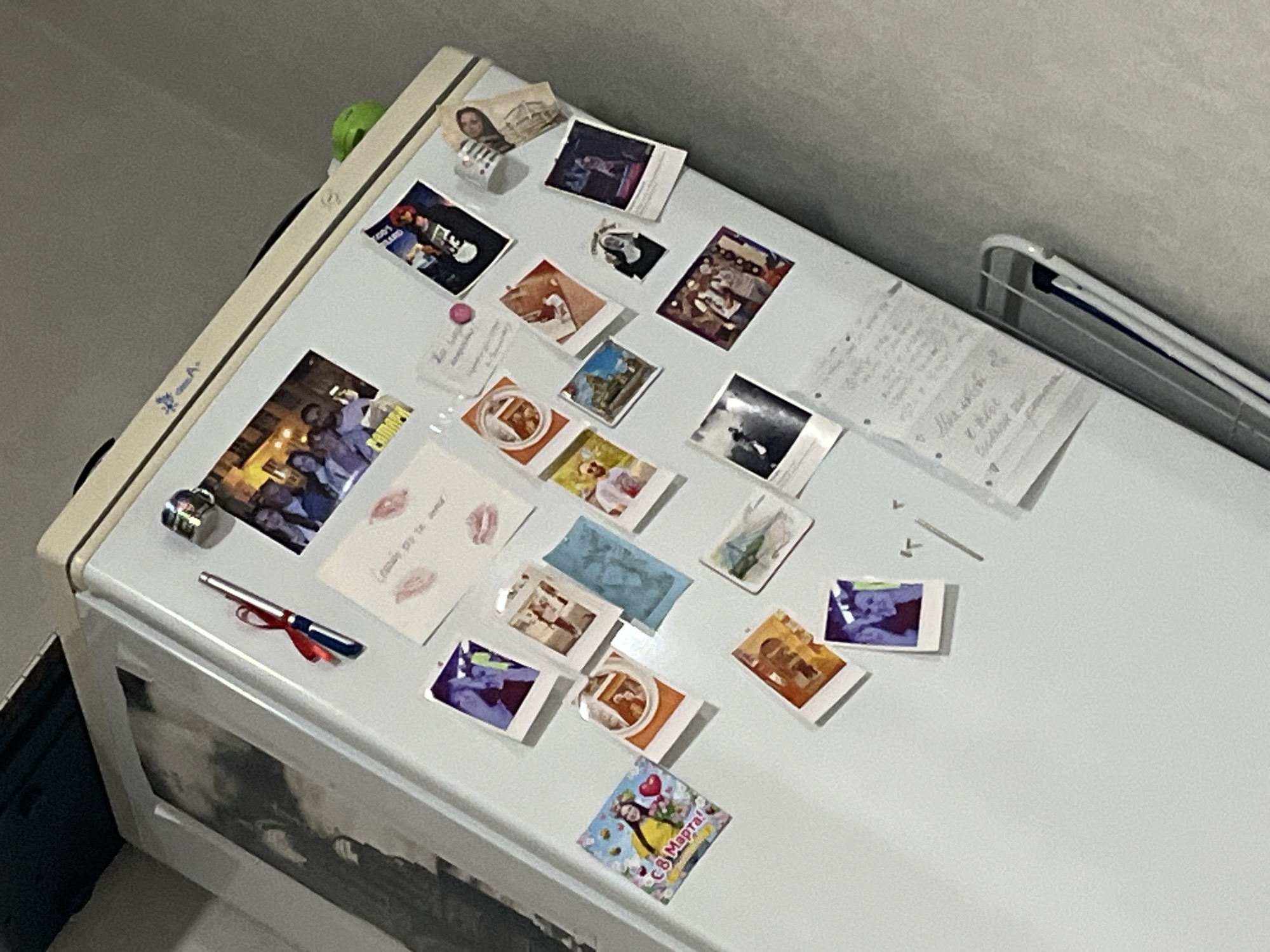 The image shows the side of a white refrigerator door, covered with various magnetic photos and notes. The door itself is white, with a cream-colored trim along the top and left edges. The background is a neutral gray wall.
The magnets and photos are arranged somewhat haphazardly across the refrigerator door. Among the items are several small square photographs, some featuring people and others appearing to be landscapes or abstract images with vibrant colors. There is a postcard with two red lip prints and some handwritten text. A pen with a red ribbon is attached to the refrigerator door with a magnet. A small silver metallic object, possibly a compact or a small container, is also visible. There is a larger black and white photo at the bottom of the refrigerator door.
One of the photos, a square with a yellow background, has Russian text that reads "8 Марта!", which translates to "March 8th!" or "International Women's Day!". This suggests the photo is a celebratory card or image related to this holiday. Another small photo shows a group of people gathered. A paper with handwritten text in what appears to be Cyrillic script is also attached to the door.
The overall impression is of a lived-in space, likely a kitchen, where personal mementos and reminders are displayed. The lighting appears to be indoor, with no clear indication of the time of day or weather. The presence of Russian text provides a strong clue to the location.