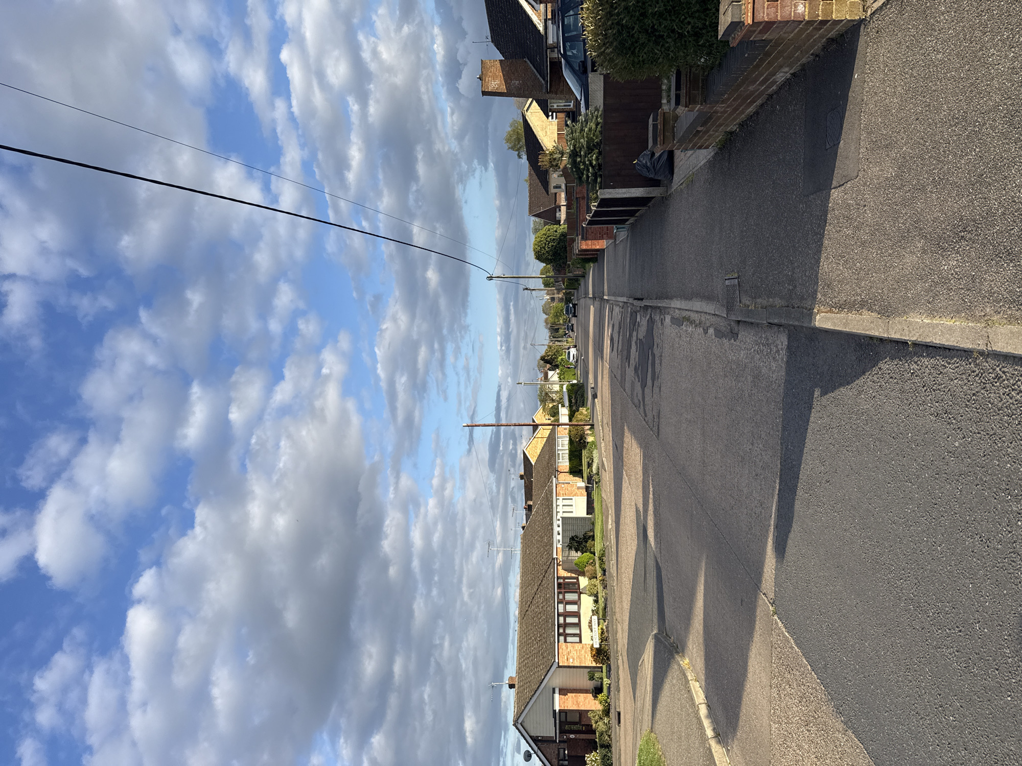 The image displays a tranquil residential street, likely situated in Colchester, United Kingdom, under a bright, partly cloudy sky. The view is down the length of the road, which extends into the distance, bordered by houses on both sides.
On the left, a row of predominantly brick and rendered houses, appearing as bungalows or low-rise dwellings with pitched roofs, lines the street. Many of these homes feature small front gardens with various shrubs and greenery. Overhead utility lines are visible, stretching across the sky between poles that punctuate the streetscape. A dark-colored vehicle is parked further down the road on this side. On the right side of the street, the paved sidewalk and the immediate fronts of properties, including fences and green planting, are in view. A black refuse bag rests on the pavement near a boundary.
The asphalt road shows some patching but appears generally well-maintained. Long, distinct shadows are cast across the road and pavements from the houses and utility poles, indicating that the sun is low in the sky, suggesting either early morning or late afternoon. The sky is a blend of clear blue patches and numerous fluffy white and grey cumulus clouds.
No people or specific activities are visible, contributing to the calm and everyday atmosphere of the scene. There is no discernible text legible on any signs, vehicles, or house features within the frame.