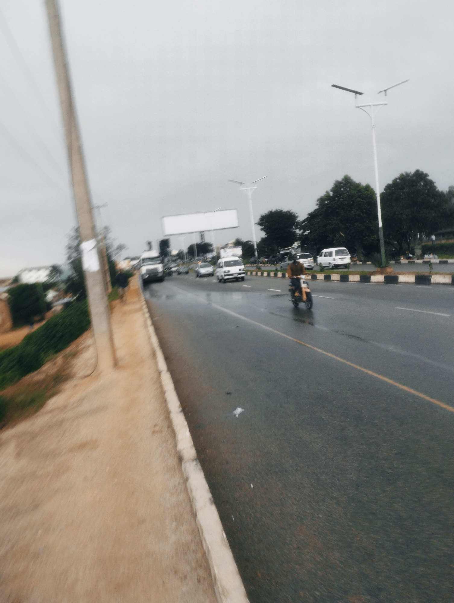 The image depicts a street scene in Jos, Nigeria. A paved road with a yellow lane marker runs through the frame, wet from recent rain. Several vehicles are present, including cars, a truck, and a white van. A motorcycle with a rider is in the foreground, moving along the road. On the left side of the road, there is a dirt path, a concrete curb, and a utility pole. Lush green vegetation and trees line the opposite side of the road, interspersed with streetlights. In the background, buildings and more trees are visible under an overcast sky.