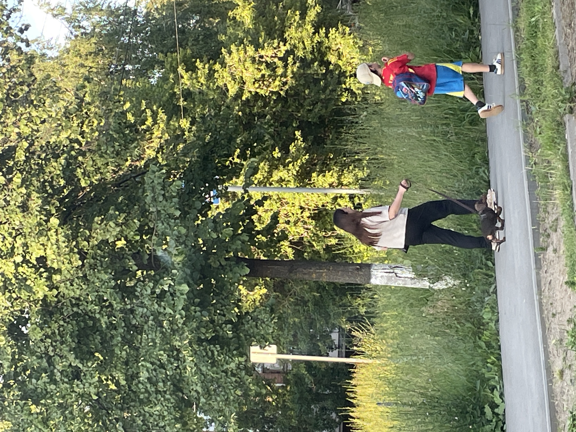 In Votkinsk, Russia, on a sunny day, a young woman walks a small dark-colored dog on a leash along a paved sidewalk. She wears a light-colored shirt and dark pants. A young boy in a red shirt, blue shorts, and a beige cap walks ahead of her, carrying a backpack adorned with colorful patches. They are surrounded by lush green foliage and tall grass. The scene is framed by large trees and a clear sky.
