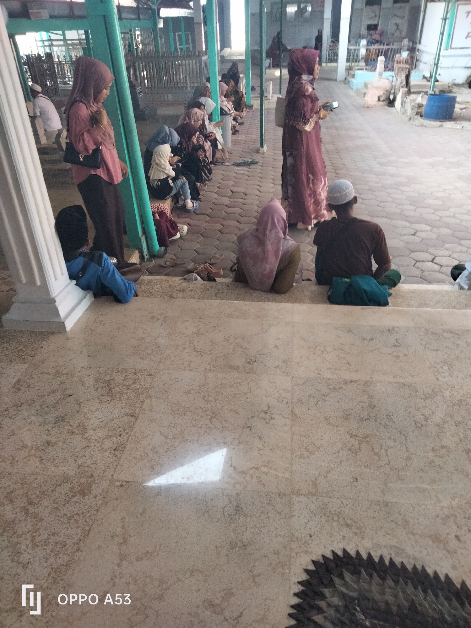 In Badung, Indonesia, a group of people, predominantly women and a few men, are seen gathered in an outdoor area with a tiled floor. The setting appears to be a place of worship or a significant cultural site, possibly a cemetery, given the presence of what looks like headstones and a blue plastic barrel in the background.
Several individuals are seated on steps or the ground, some engaged in prayer or contemplation. A woman in a light pink headscarf is standing, holding a black purse and a mobile phone. Another woman in a long, patterned dark dress is also standing, holding a phone. A young person wearing a blue jacket is sitting on the ground leaning against a white pillar. A man in a brown shirt and white cap is seated on the steps, with a green cloth and a teal bag beside him. Other individuals are visible in the background, some in traditional attire, indicating a cultural or religious gathering. The overall atmosphere suggests a serene and respectful gathering.