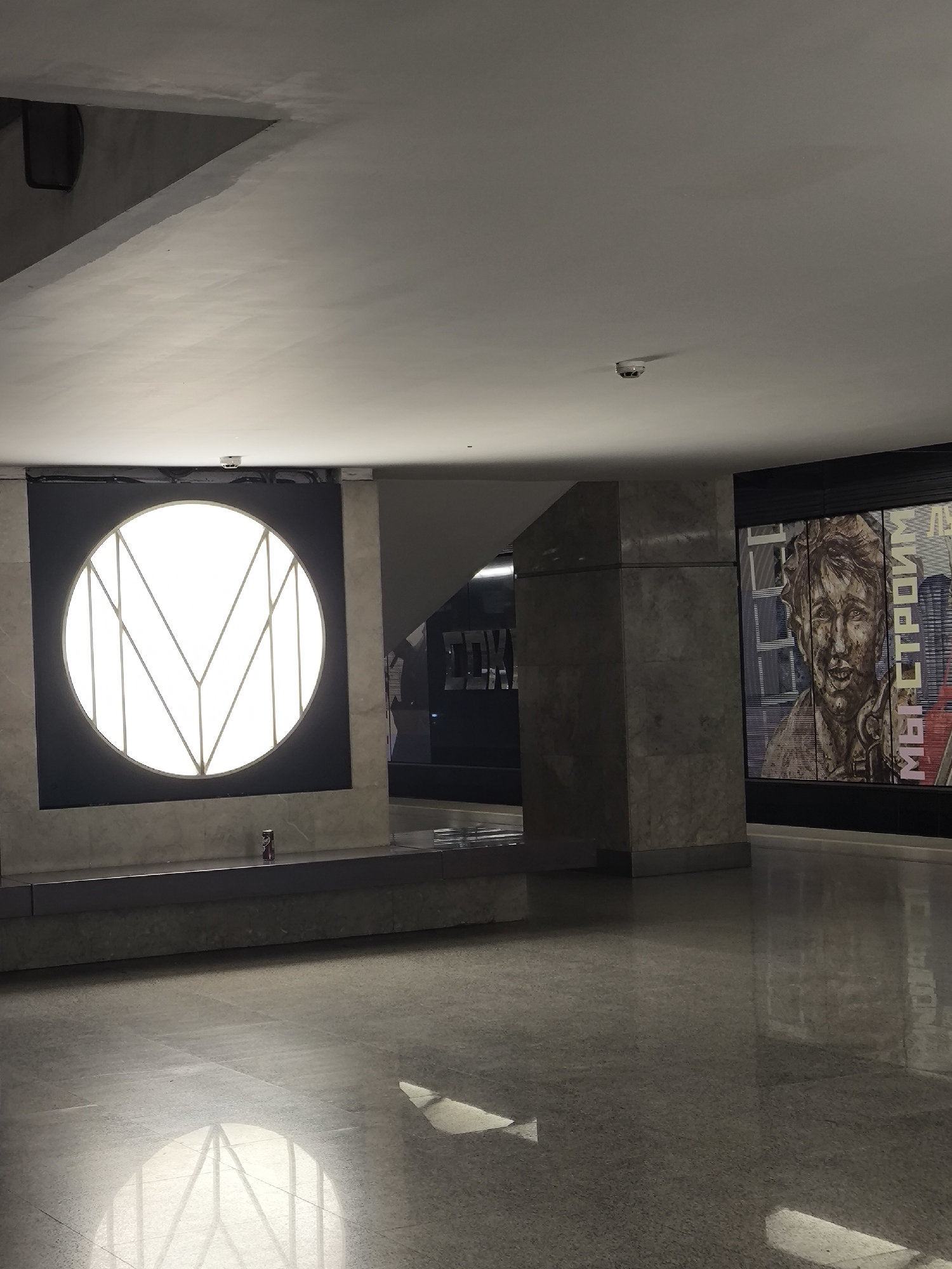 The image depicts a spacious indoor area, likely a metro station in an unknown city. The floor is a shiny, grey marble, reflecting light from a large, circular light fixture on the left. The light fixture is set against a dark background and appears to have a stylized "M" design. In the background to the right, there is a large mural with a portrait of a man and some text. There is a security camera on the ceiling. There is a can of soda on a bench in front of the light fixture. The setting appears to be empty.