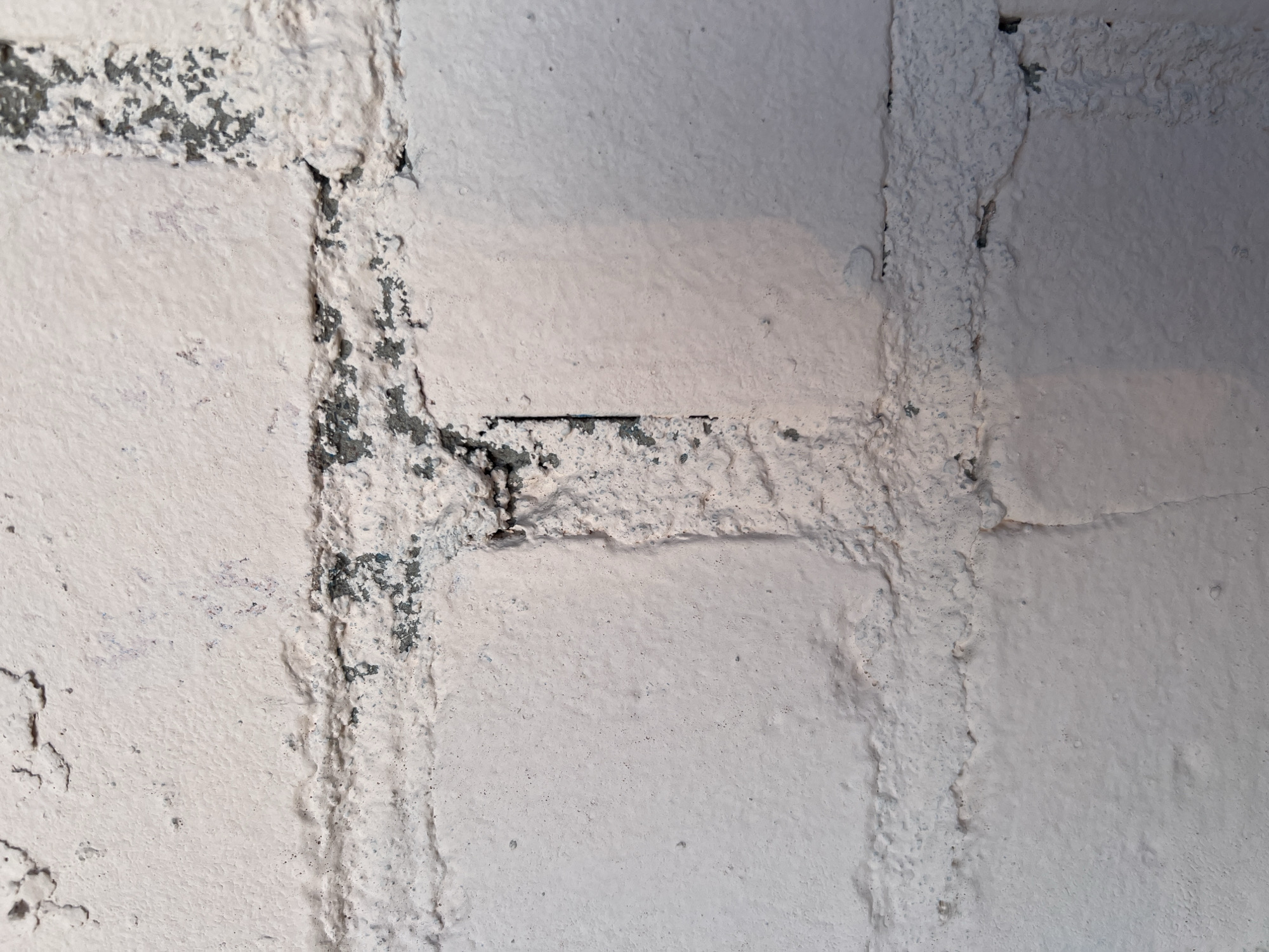 The image shows a close-up of a textured, white-painted brick wall. The bricks are laid in a stretcher bond pattern, with the mortar joints creating a grid-like structure. The paint is unevenly applied, with some areas showing the rough texture of the underlying brick and mortar, while other areas appear smoother. There are visible signs of wear and tear, including chipped paint and some areas where the dark brick or mortar is exposed. The lighting suggests an indoor setting, possibly an unfinished or industrial space, with soft, diffused light casting gentle shadows. No people, animals, or specific objects are visible in the frame, and there is no discernible text. The overall impression is one of an aged and weathered construction.