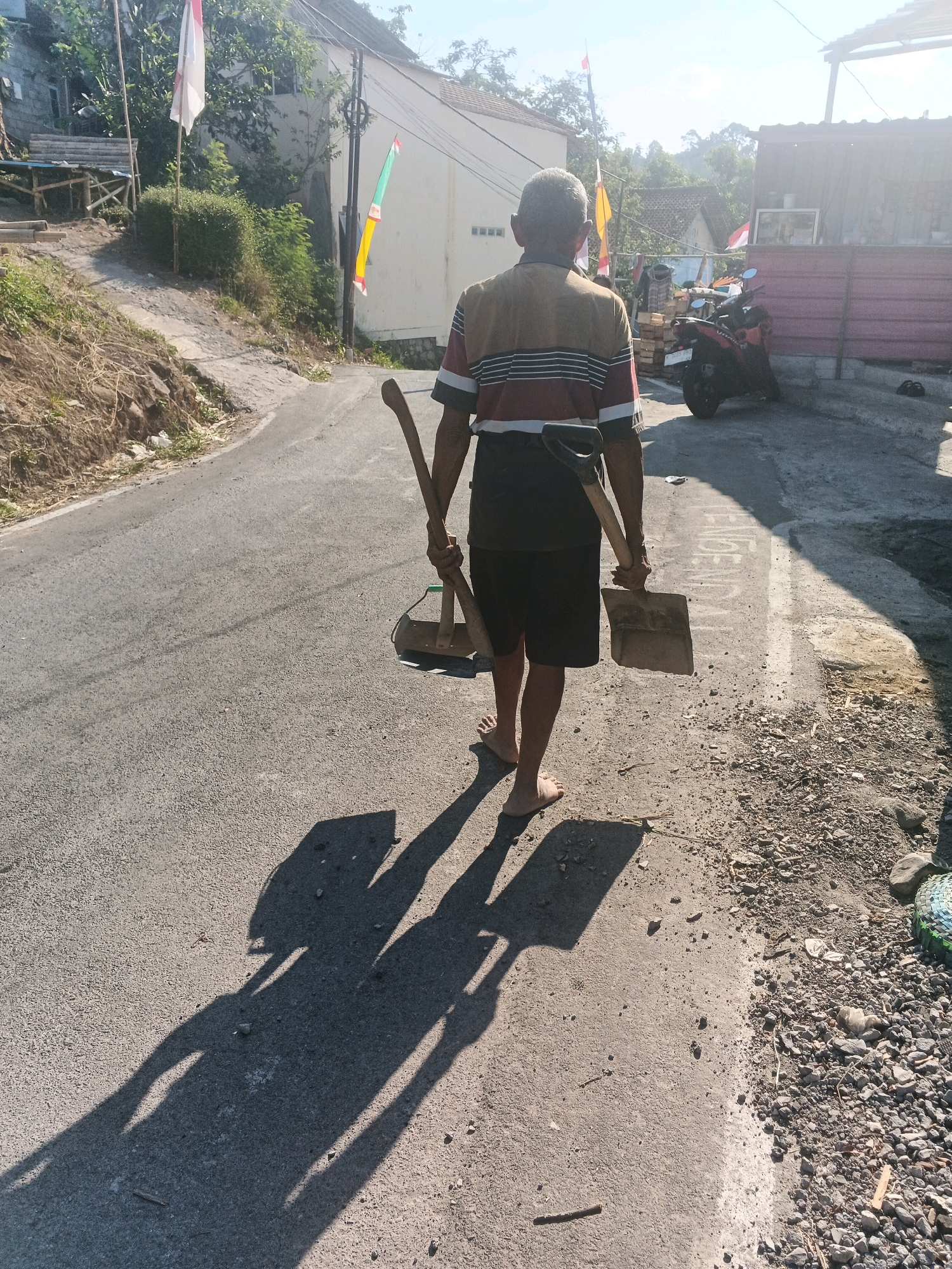 An elderly man with gray hair, wearing a striped t-shirt and dark shorts, walks away from the camera down a paved road. He carries a shovel in his right hand and an axe in his left. His feet are bare. The road appears to be in a rural or semi-rural area, possibly a village, with houses and vegetation visible in the background. The sun is shining brightly, casting long shadows from the man and his tools onto the road. The location is identified as Keji, Indonesia.