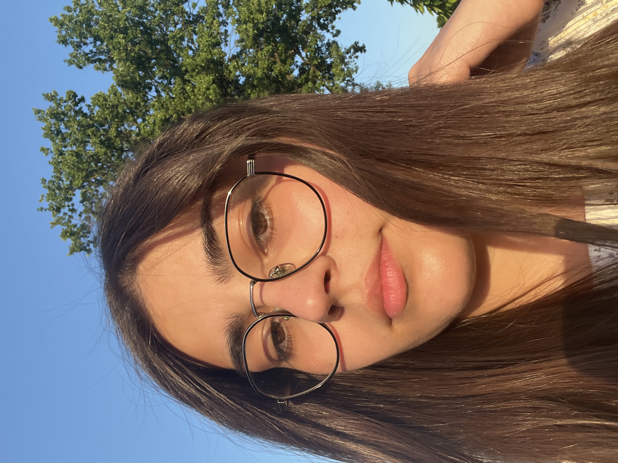 A young woman with long, dark brown hair and wearing round, wire-rimmed glasses is pictured outdoors. The golden hour sunlight casts a warm glow on her face, highlighting her features. She is looking directly at the camera with a neutral expression. Behind her, a lush green tree with dense foliage stands against a clear blue sky. The image appears to have been taken in Dubivtsi, Ukraine.