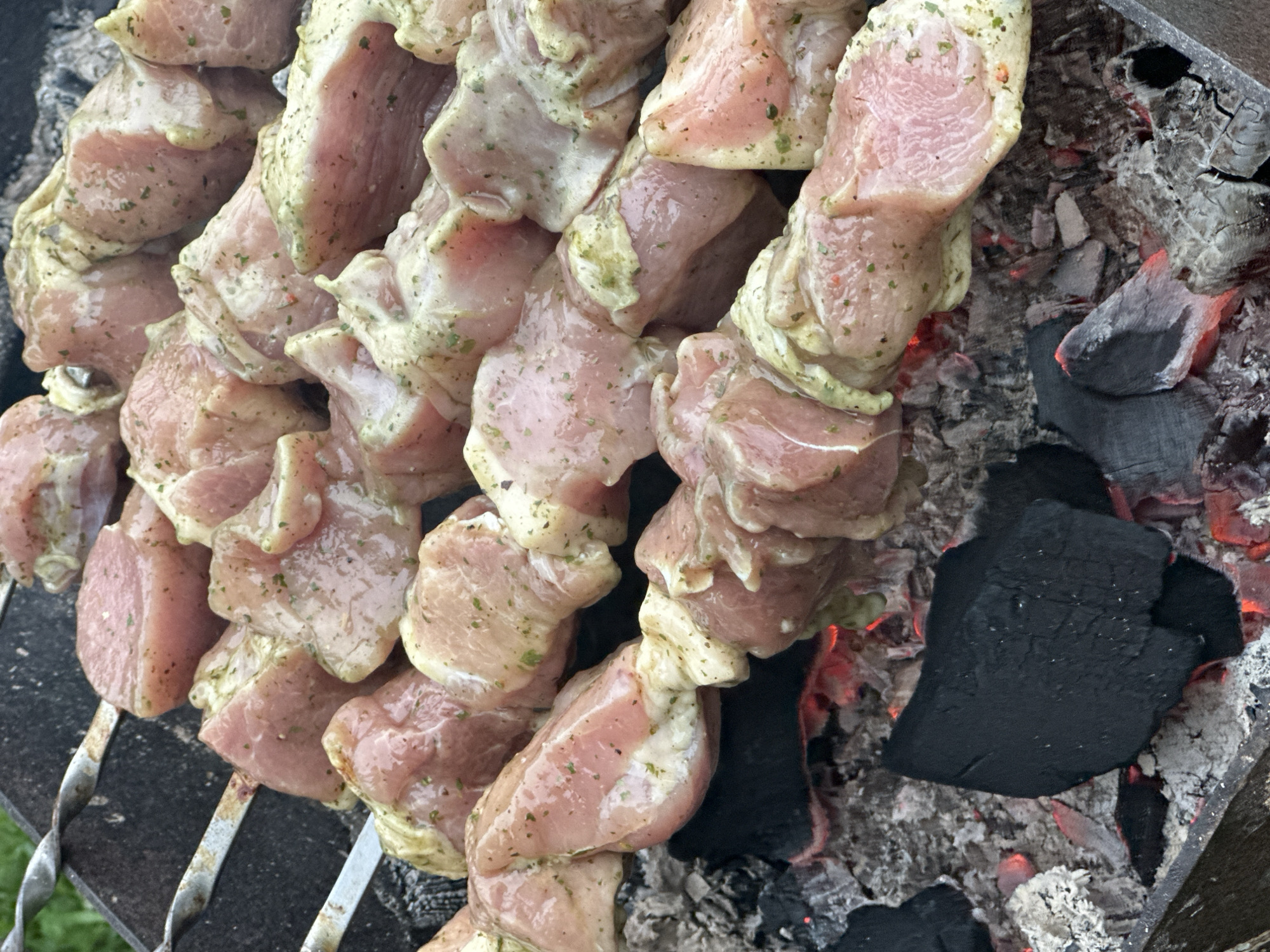 The image shows a close-up view of marinated meat skewers being grilled over hot coals. The meat is cut into bite-sized pieces and seasoned with herbs and spices. The skewers are placed on a metal grill rack, and the glowing red coals underneath indicate that the barbecue is active. The background is slightly blurred, but it appears to be an outdoor setting with greenery visible, suggesting a backyard or a picnic area. The specific location in the city of Подмой, Russia is not discernible from the image. The focus is entirely on the food being prepared.