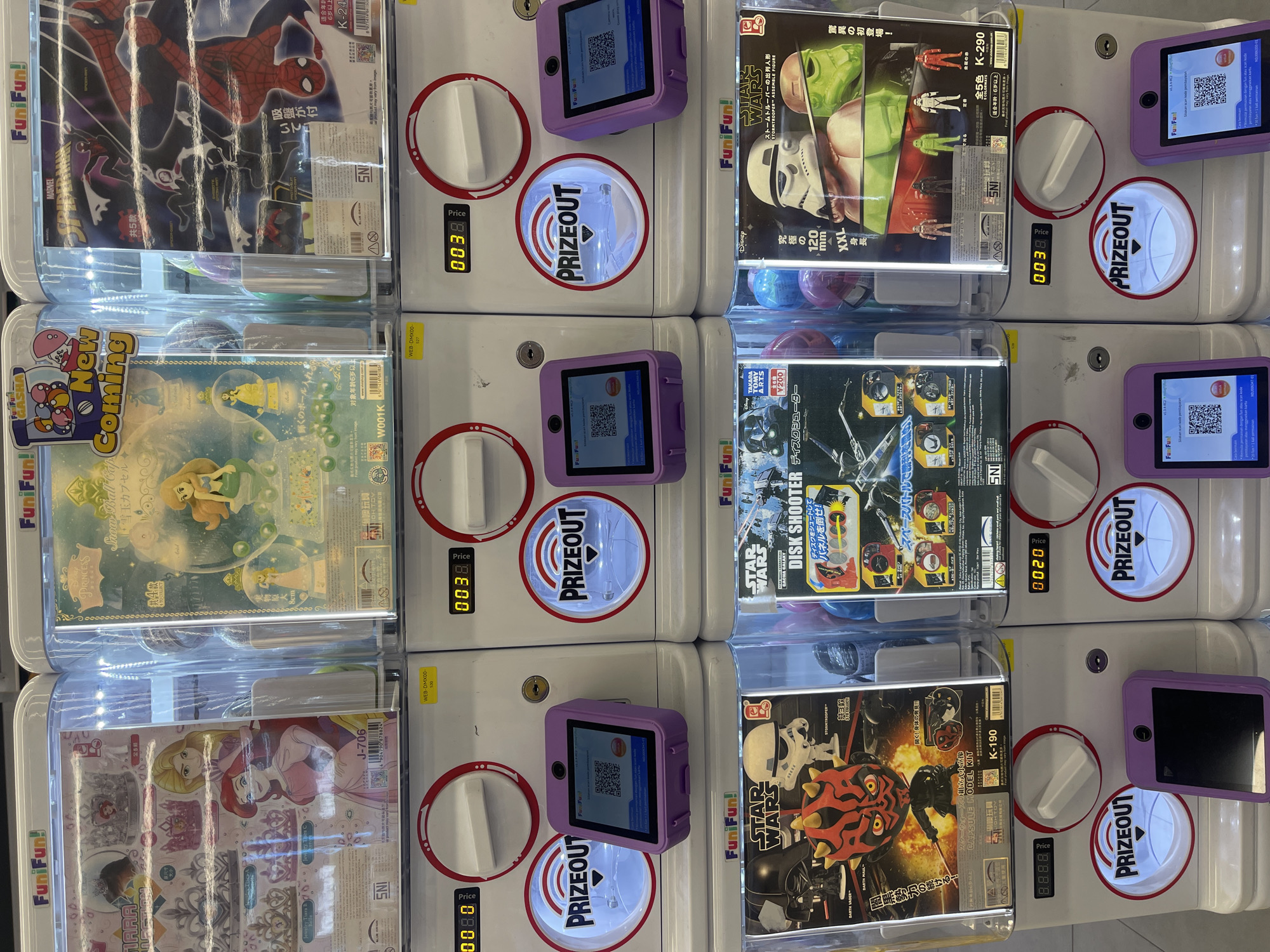 The image shows a row of capsule toy vending machines, likely found in a shopping mall or entertainment venue. The machines are branded "FunFun" and feature popular franchises like Disney Princesses, Marvel's Spider-Man, and Star Wars. Each machine has a display window showcasing the available toys, a slot for payment, and a "Prizeout" chute. Some machines also have small purple screens with QR codes, possibly for payment or app integration. The overall environment appears clean and brightly lit, suggesting an indoor retail space. The presence of Japanese text and branding indicates that these are likely imported goods. The machines are arranged in a grid, and the scene is captured from a slightly elevated, head-on perspective. The backdrop is out of focus, but hints of other retail displays are visible. There are no people visible in the image. The vending machines are the primary focus.