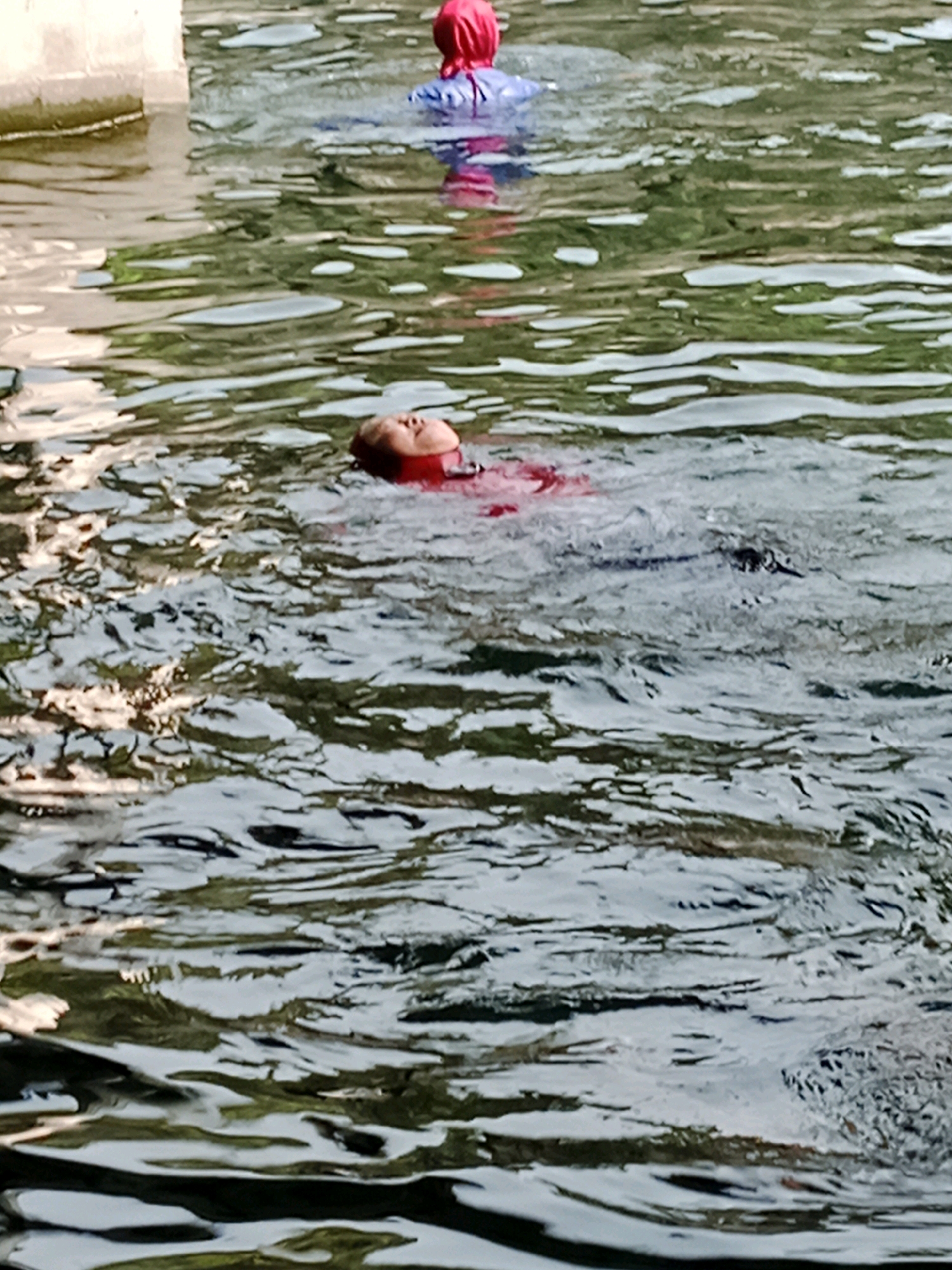 In Jentekan, Indonesia, a person is floating on their back in the water, their face visible above the surface. They are wearing a red garment and a blue buoyancy aid. Another person is partially submerged in the water further in the background, also wearing a red head covering and blue and pink clothing. The water is murky and reflects the surrounding environment, with ripples disturbing the surface. A concrete structure is visible on the left edge of the frame.