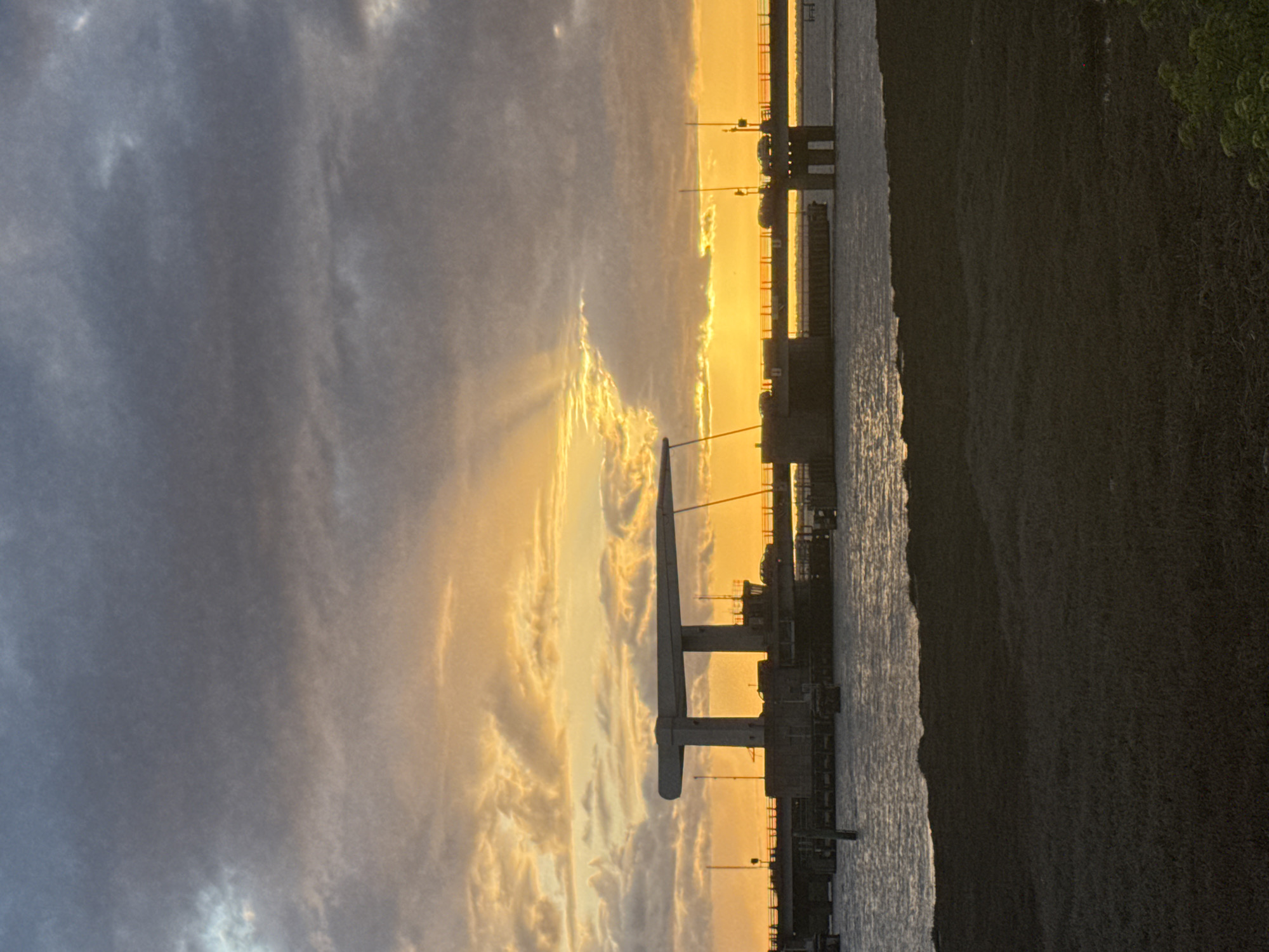 The image shows a sunset view of the North River pier in Great Yarmouth, United Kingdom. The pier structure is silhouetted against the bright orange and yellow sky, with dark clouds on the left side of the image. The water reflects the colors of the sunset. On the right side of the image, there is a dark landmass, presumably the shore, with some greenery visible. Streetlights are seen along the pier.