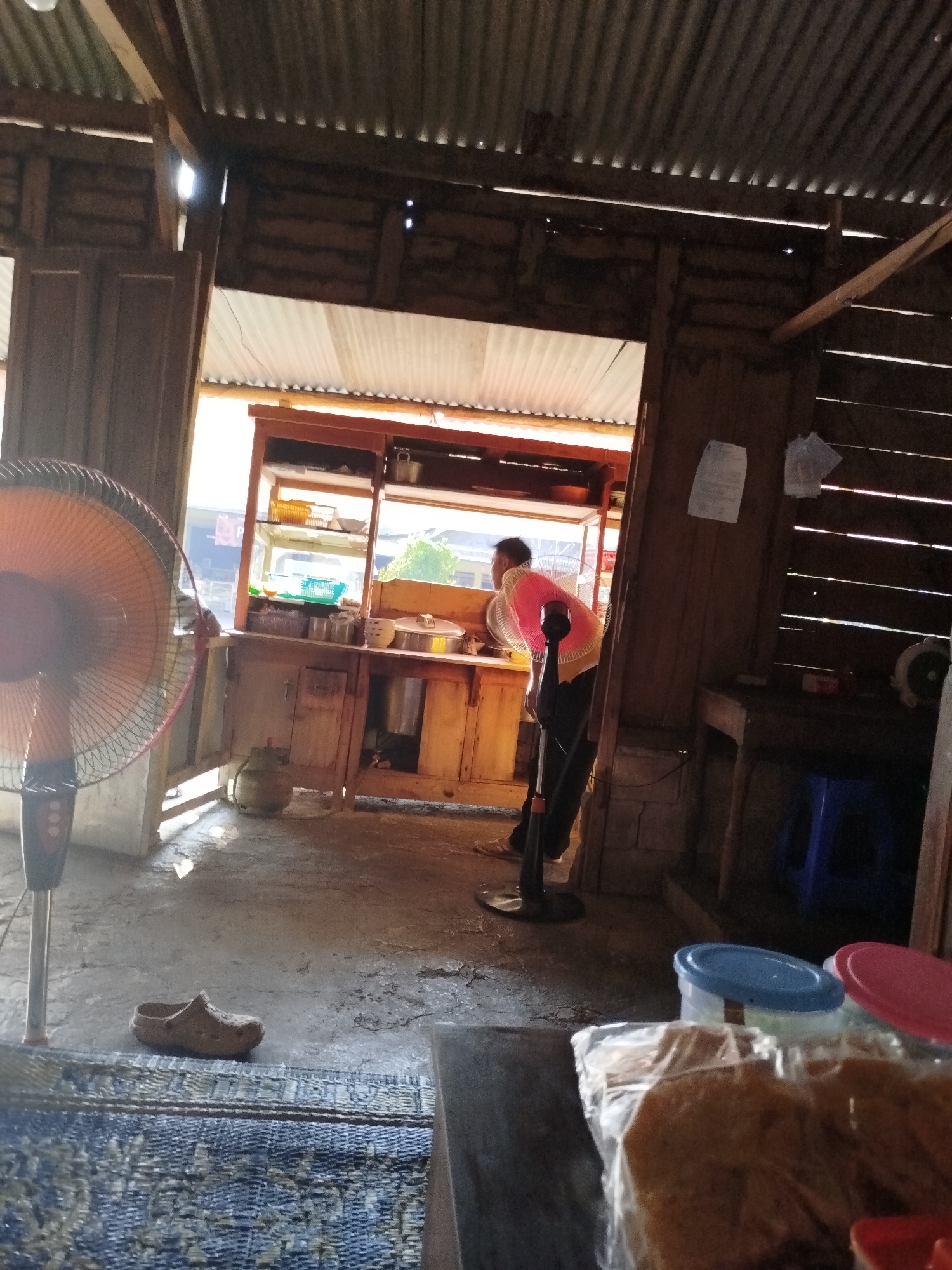 The image depicts an indoor scene, likely a shop or food stall, with a rustic wooden interior. A person, wearing a pink top and dark pants, is visible in the background, standing near a counter with shelves displaying various items. A large standing fan with an orange grille is prominently placed in the left foreground. The floor appears to be concrete, with a woven mat laid out in the lower left. A single brown shoe is on the floor near the mat. The overall lighting suggests it is daytime, with sunlight entering from an opening or window. The environment suggests a casual, possibly rural, setting within Pojok, Indonesia.