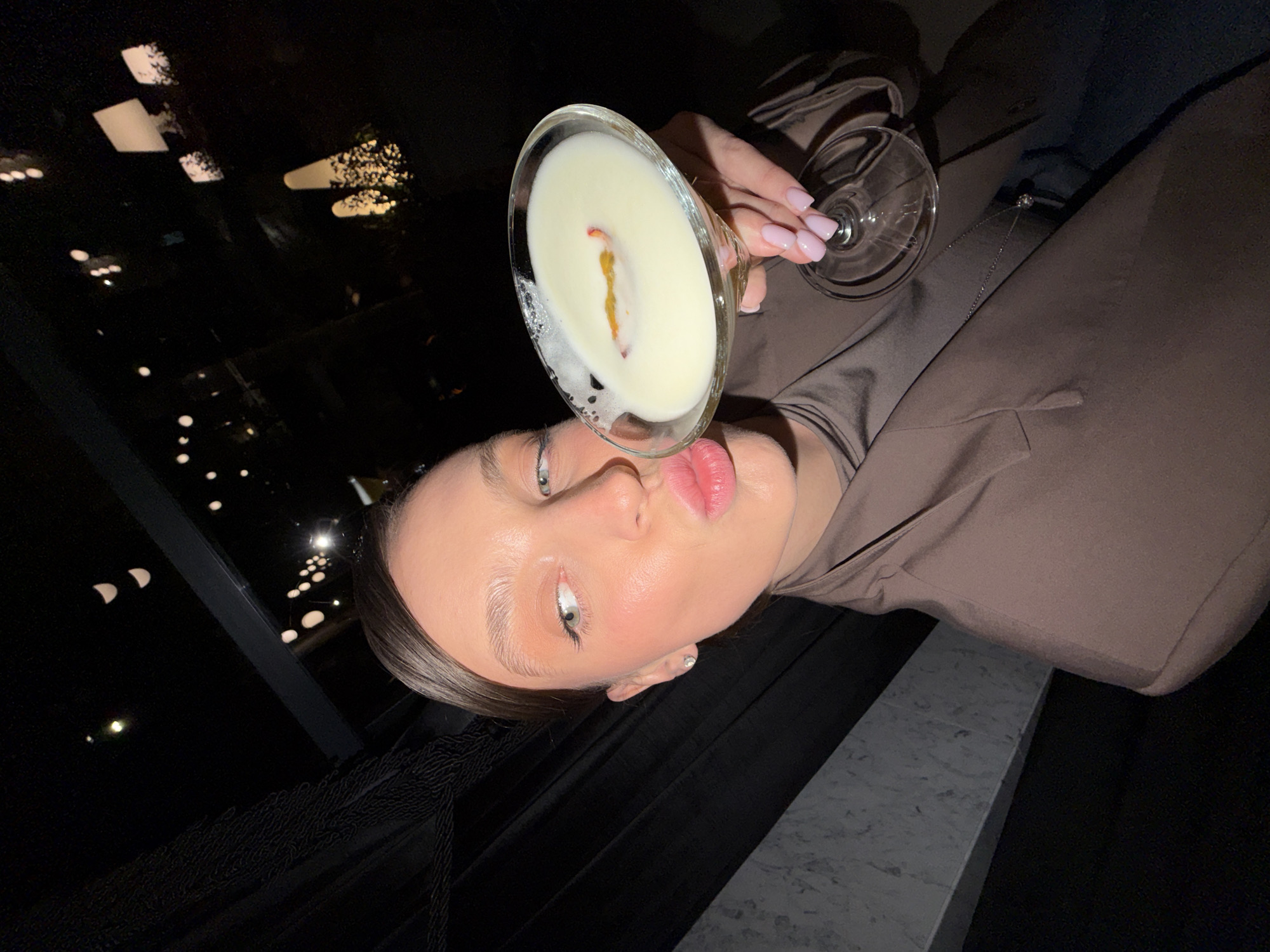 A person is holding a cocktail in a martini glass in what appears to be a restaurant or bar setting. The drink has a creamy white appearance with a piece of garnish floating on top. The background is dark, with abstract lighting fixtures visible through a window. There is black curtain on the left side. The person is wearing a brown dress or jacket and a silver necklace. The location could be Budva, Montenegro, but further location data is needed to confirm.