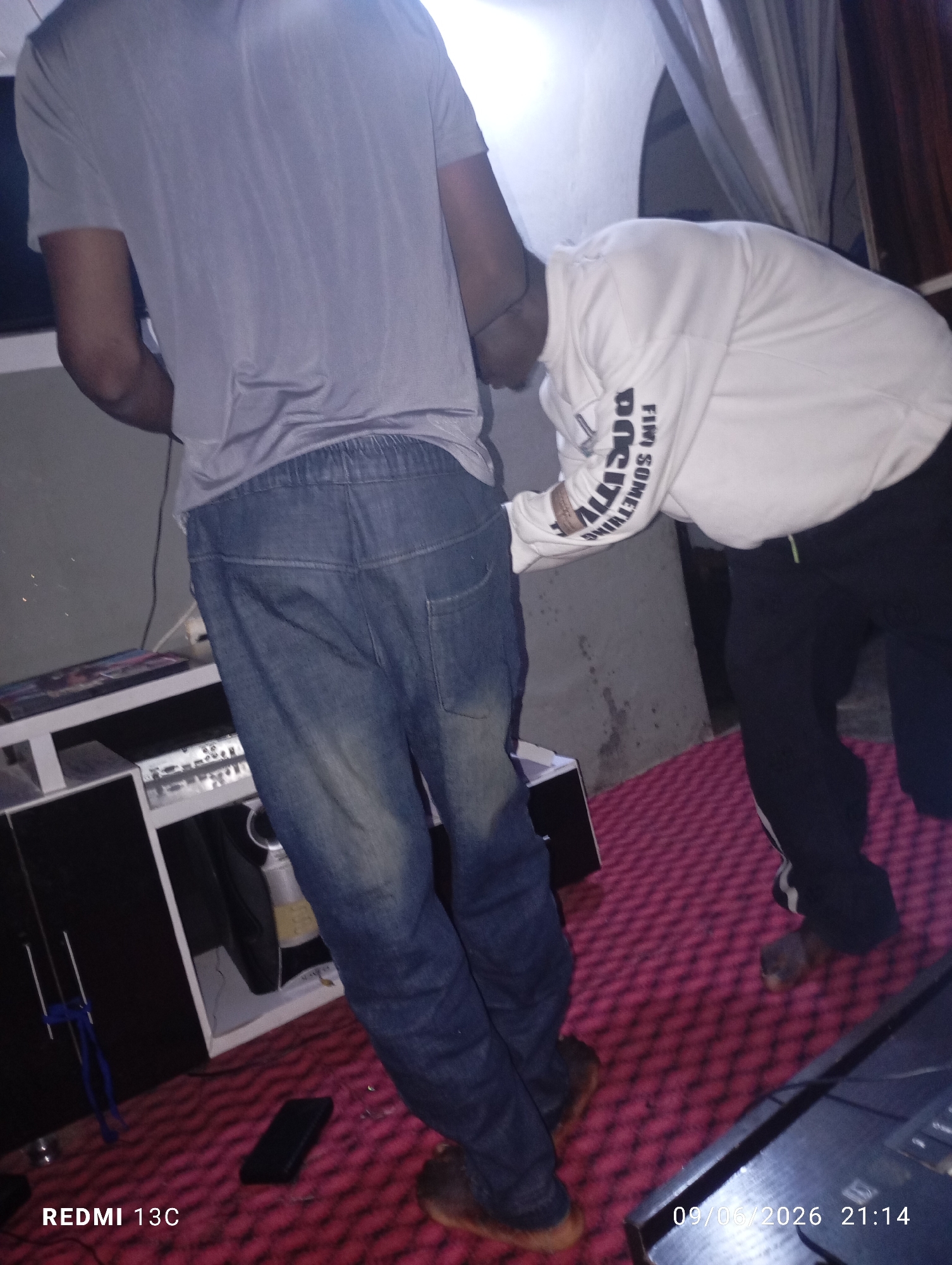 Two individuals are standing in an indoor environment. The person on the left is wearing a grey t-shirt and blue jeans. The person on the right is wearing a white hoodie with black lettering on the sleeves and black pants. The floor is covered with a pink textured rug. In the background, there is a white shelf unit with electronic equipment. The scene appears to be dimly lit, possibly a residential area.