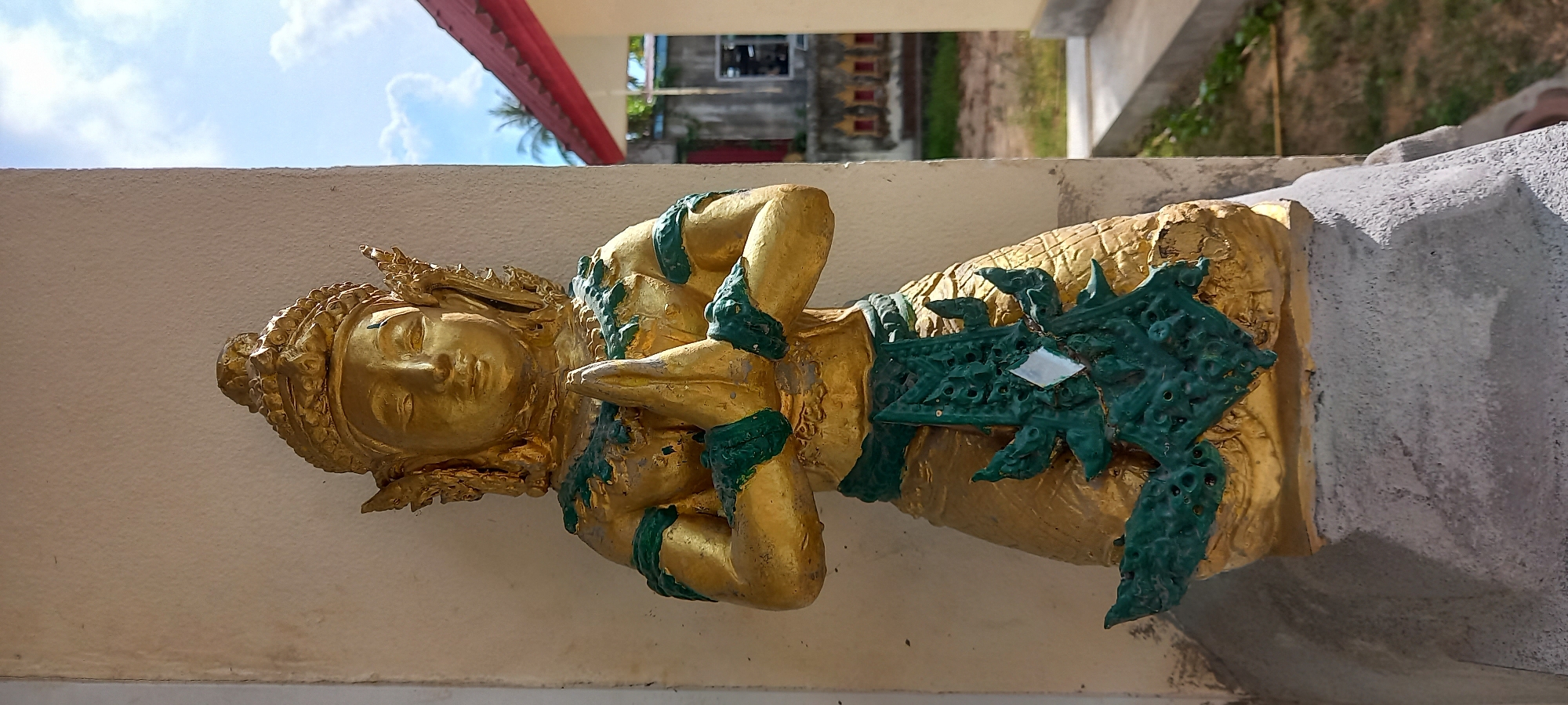 A horizontal, medium shot shows a golden statue of a figure with green ornamentation, mounted on a white wall. The statue depicts a person with eyes closed and hands crossed over their chest. They are adorned with a headdress and multiple green bands on their arms and waist. The wall continues to the right of the statue, ending in a corner. Behind the wall and statue, there is a blurred view of a street with trees, a building, and a partly cloudy sky. The statue is in Moo 4, Thailand.