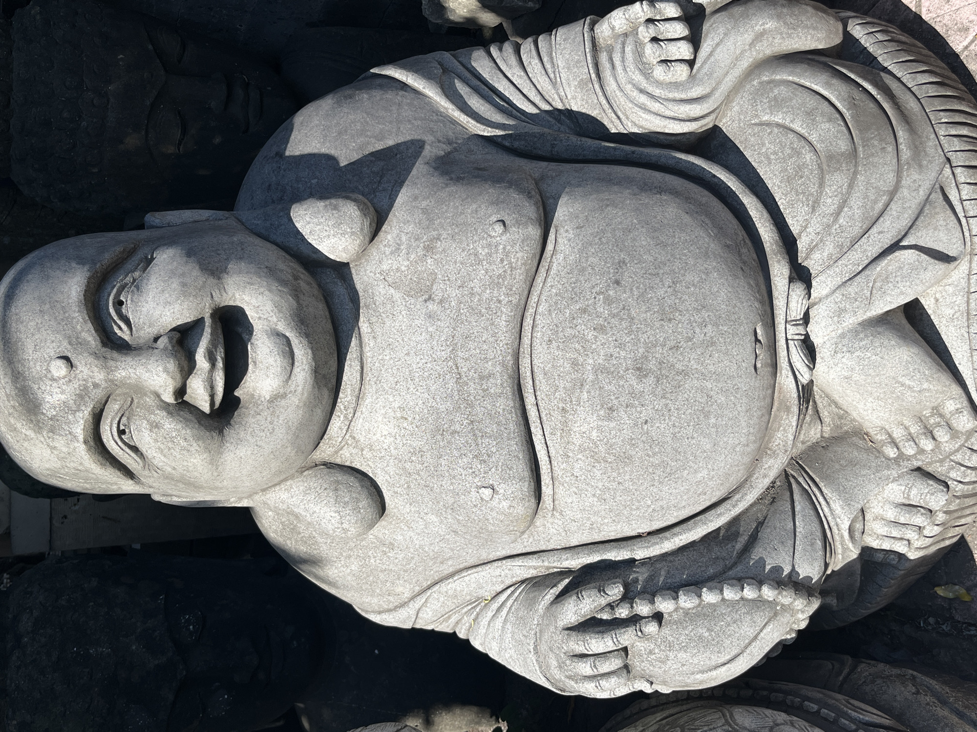 A stone statue of a laughing Buddha, or Budai, sits in the foreground. The statue is carved from light-colored stone, with a weathered texture. The figure is depicted as rotund, with a smiling, jovial expression, wide eyes, and a round face. The Buddha's hands rest in his lap, one holding a string of prayer beads. The statue is seated, with legs crossed and draped in stylized robes. The background is dark, with the faint outlines of other stone statues visible, suggesting a marketplace or a display of religious artifacts. One of these background figures appears to be a darker stone statue of a meditating Buddha. The ground beneath the statue is visible, appearing to be an outdoor setting with some fallen leaves. The lighting suggests it is daytime, with sunlight casting shadows. This scene is located in Sukawati, Indonesia, a region known for its art markets and craftmanship.