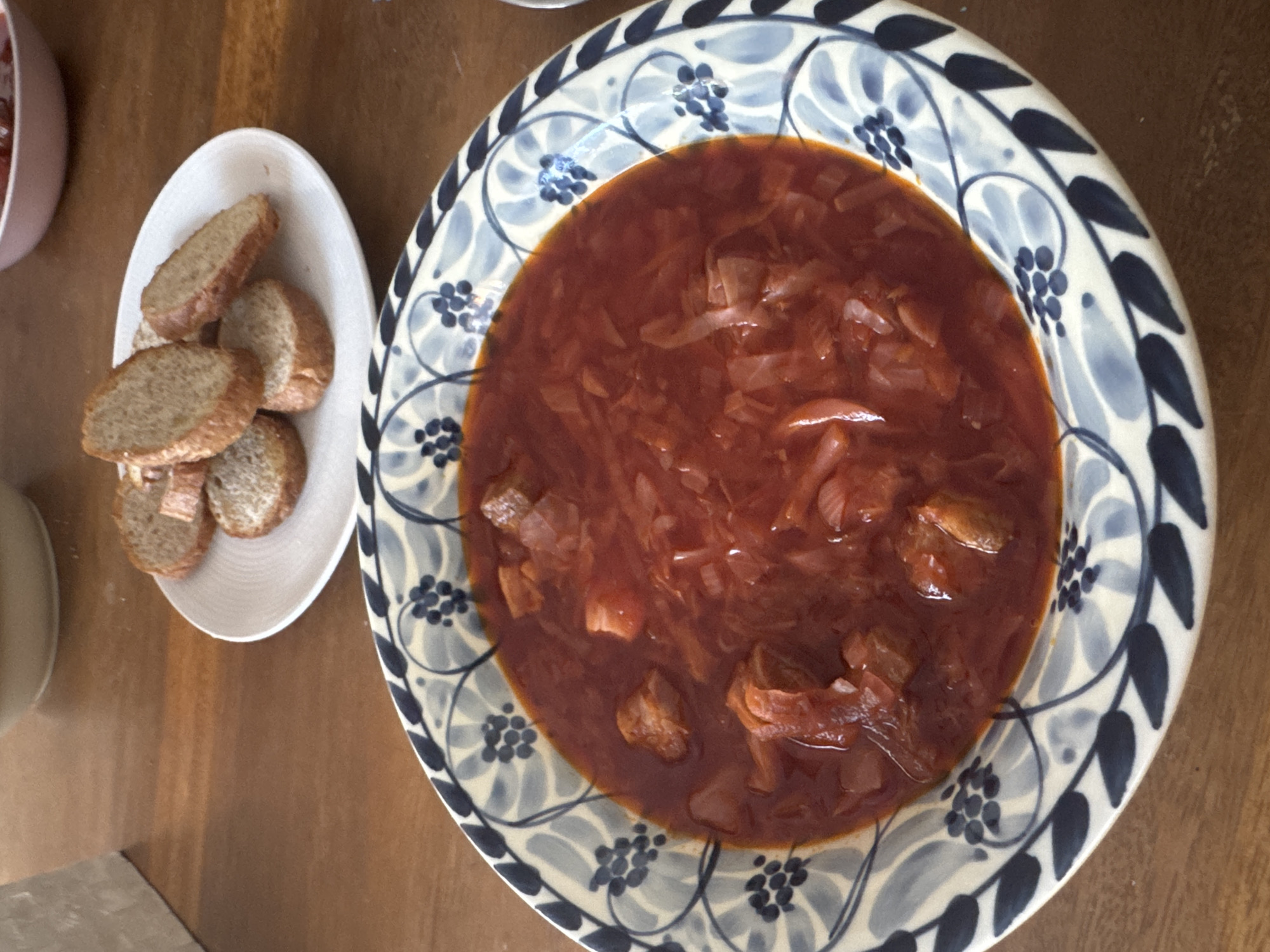 A high-angle shot shows a wooden surface with a plate of soup and a plate of bread. The soup is a dark red color with visible pieces of meat and cabbage, served in a round, decorative bowl with a blue and white floral pattern around the rim. Next to the bowl is an oval plate containing several slices of bread. Parts of a pink bowl and a beige bowl are visible on the left edge of the frame. The location and city details provided, Kuta Selatan, Indonesia, are not discernible from the image.