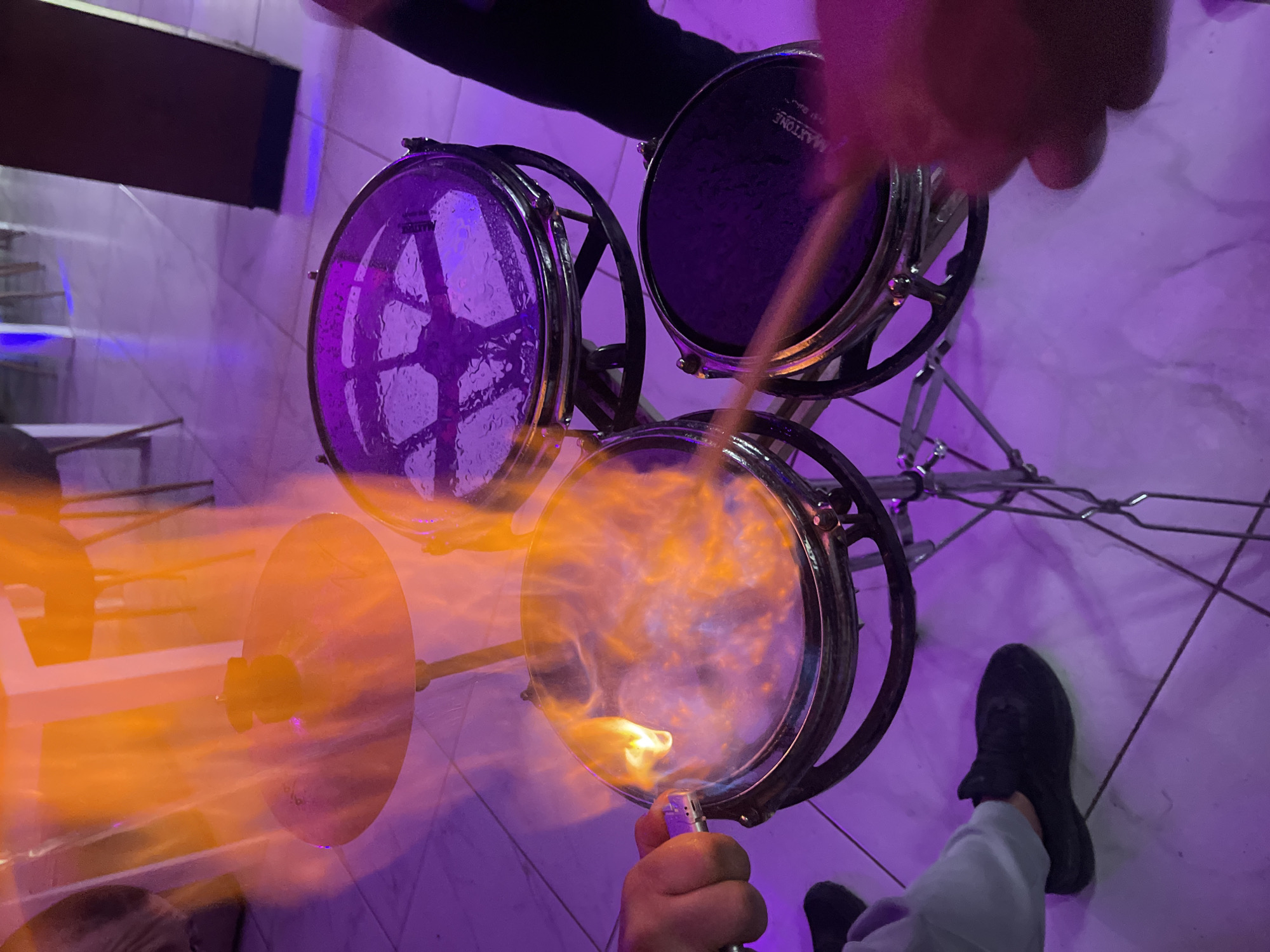 A view from above showing three small drums on a stand, with a cymbal to the left and a lighter igniting the bottom drum. The drums are metal with black drum heads except for the ignited one. A drumstick is held above the drums. A person’s legs and feet are visible at the bottom. The scene appears to be indoors, illuminated with purple light. It is impossible to confirm the location in Somovit, Bulgaria, based on this image.