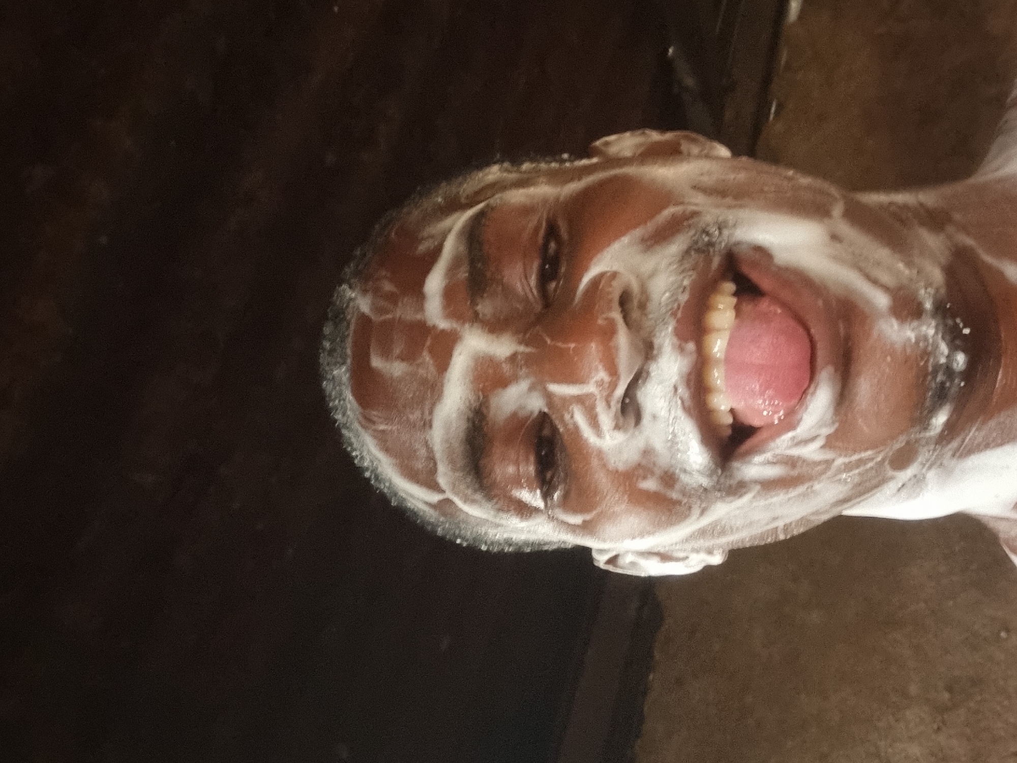 The media file displays a close-up, rotated portrait of an adult's face almost entirely covered in white soap lather or foam. The person, appearing to be male with short, dark hair visible through the foam, is looking directly at the camera with an open mouth, revealing a full set of teeth and a pink tongue. A wide smile or laugh is evident, conveying a sense of joy, amusement, or playfulness. The eyes are open and visible amidst the foam. The activity depicted is consistent with someone washing their face or head, possibly in a shower or a bathroom. The background is dark and largely indistinct, suggesting an indoor setting with low light or dark-colored surfaces. No visible text is present in the image. The location context provided is Onitsha, Nigeria.