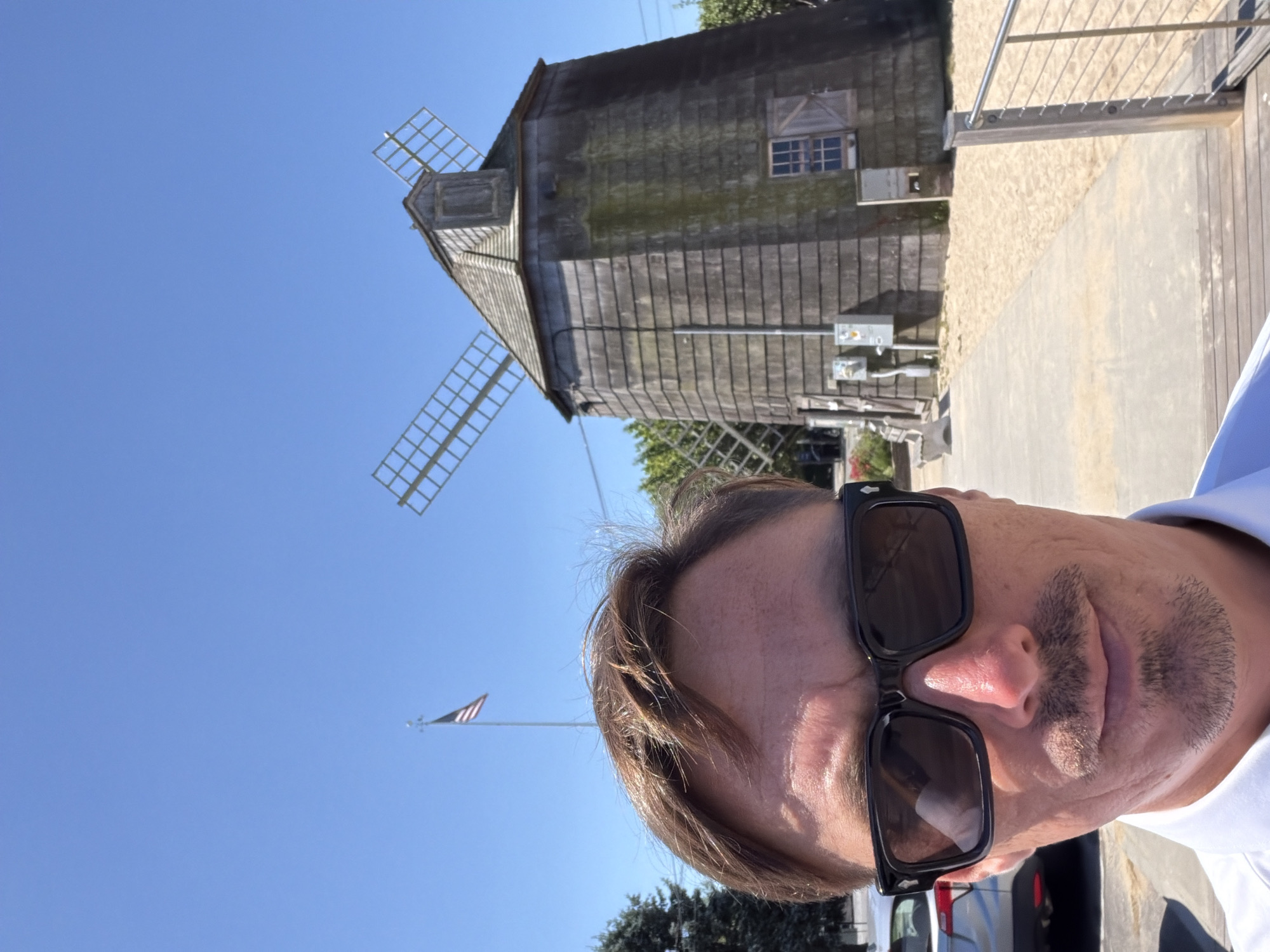 A man wearing sunglasses and a white shirt is taking a selfie in front of a wooden windmill. The windmill has four blades, two of which are visible and are made of a light-colored material. An American flag is flying on a flagpole to the left of the windmill. The ground is covered in sand, and a paved walkway leads to the windmill. In the background, there is a silver car parked to the left of the frame, and some trees and greenery. The sky is clear and blue. The scene appears to be outdoors on a sunny day in the Village of Sag Harbor, United States of America.