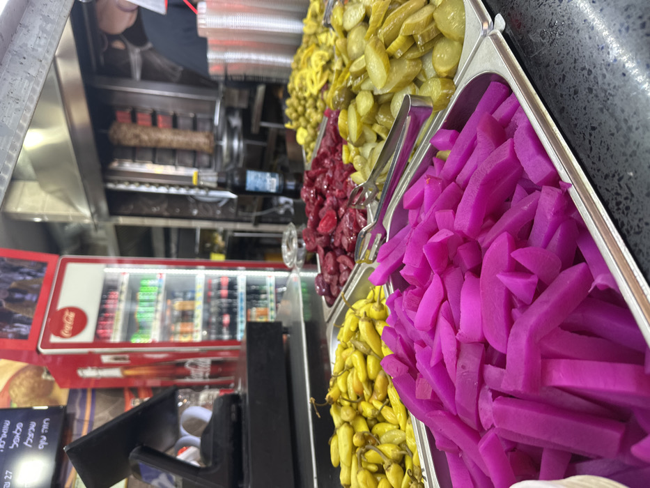The image depicts the interior of a food service establishment in Mishmar Hasharon, Israel, focusing on a counter display of pickled items. In the foreground, a black countertop holds four rectangular stainless steel trays. From left to right, these trays contain bright magenta-colored pickled daikon radish cut into sticks, yellow pickled small chili peppers, dark red pickled beet slices, and sliced green pickled cucumbers. Two sets of stainless steel serving tongs are present among the pickled vegetables.
In the midground, a black point-of-sale system is situated on the left, adjacent to a menu board displaying Hebrew text. Behind the counter, a red Coca-Cola branded upright refrigerator, fully stocked with various bottled and canned beverages, is visible. Above the refrigerator, a small screen or sign is present. To the right, a vertical rotisserie machine is actively cooking a large skewer of meat, consistent with shawarma. A dark glass bottle with a yellow cap and a stack of silver disposable cups are positioned near the rotisserie. A person wearing a dark shirt is partially visible in the background on the right side of the rotisserie. The environment suggests an active commercial kitchen and customer service area.