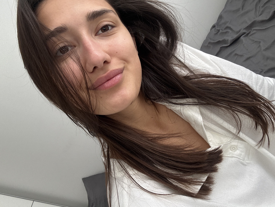 A female individual, appearing to be an adult, is depicted in a medium close-up, looking directly towards the camera with a slight smile. Her head is tilted approximately 10-15 degrees to her right. She has long, dark brown hair, which is parted slightly off-center and falls over her shoulders, with some strands partially obscuring her forehead and left eye. She is wearing a white collared shirt, possibly a polo, with two visible buttons on the placket. Her skin appears clear. The background suggests an indoor setting, likely a bedroom. To the upper left, a plain, light-colored wall is visible. Resting against this wall is a dark grey pillow. To the right and bottom, a dark grey, rumpled fabric, consistent with a bedsheet or blanket, covers a surface. The lighting is soft and even. The image contains no visual elements, landmarks, or specific architectural details to identify its location as Budva, Montenegro, or any specific address within that city; the environment appears to be a generic residential interior.