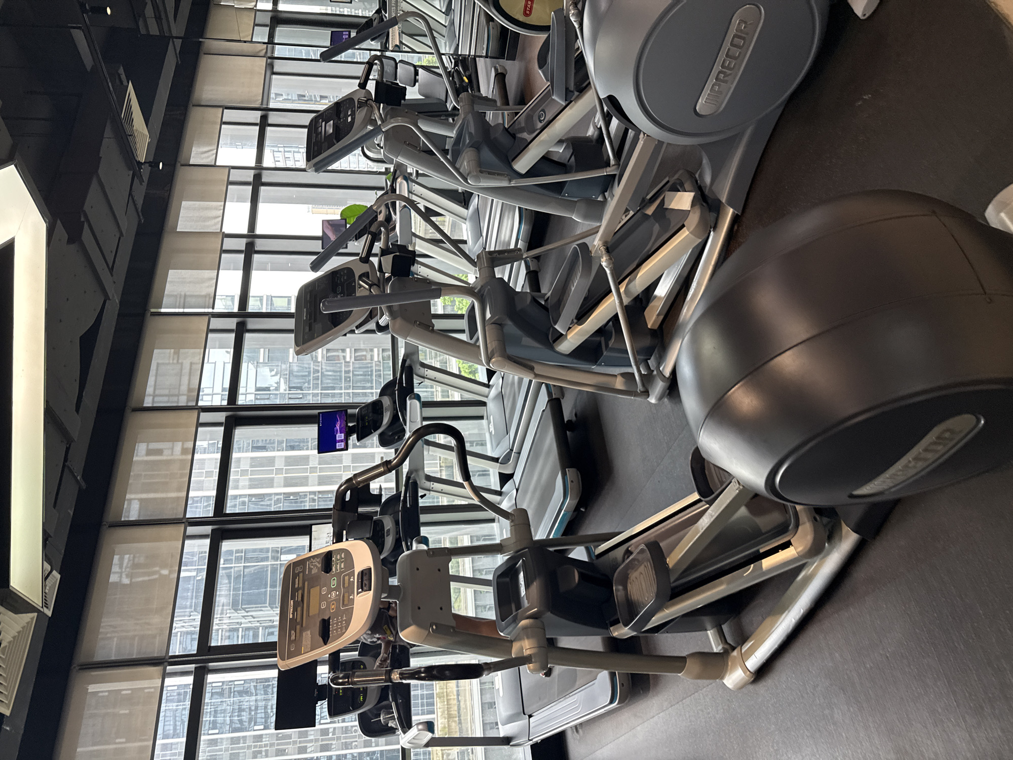 The image captures the interior of a modern gym located in Haizhu District, China. The gym features a row of cardio machines, including ellipticals and treadmills, positioned in front of large windows that offer a view of urban high-rise buildings. The machines are gray and silver, with black accents and consoles displaying various workout metrics. The floor is a dark, textured material. The lighting is bright, with ceiling fixtures visible. There are no people present in the image, and the machines appear to be idle. The overall atmosphere is clean and well-maintained, typical of a commercial fitness facility.