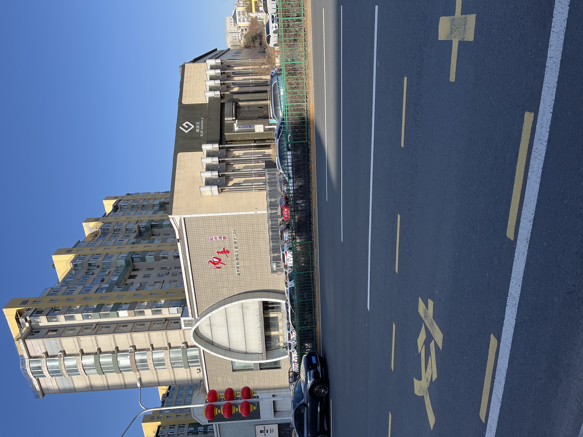 The photograph, taken in daylight, showcases a cityscape, likely in Changchun City, China, given the language on the buildings. Buildings dominate the left portion of the image, including a high-rise apartment complex and a structure with the branding "KYOSHIDAK SPRING," underneath of which is the text "通州搓背影院足道泰式SPA," and above which is the text "一私沐汤泉一" and the text "飛訊美 GRAMMY" on another part of the building. The street on the right side of the frame is marked with traffic lines. A green fence runs along the sidewalk. A line of vehicles is parked along the curb. A traffic light is visible on the bottom left. The sky is a clear blue.