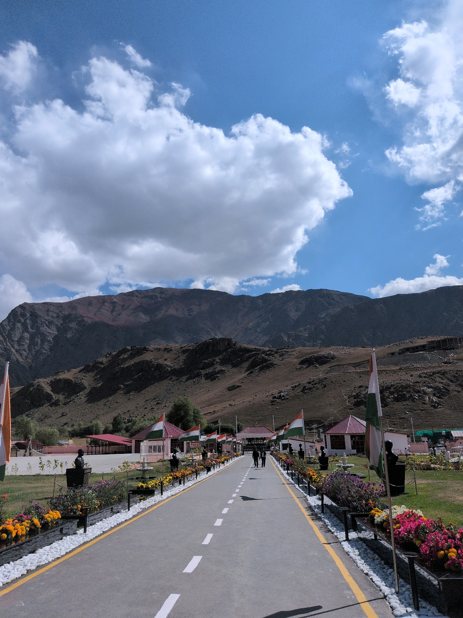 The image depicts a wide, paved road lined with a yellow stripe and white dashed lane markers, leading towards a mountainous landscape under a bright blue sky with scattered white clouds. The road is flanked on both sides by well-maintained gardens featuring rows of colorful flowers, primarily in shades of orange, yellow, pink, and purple, bordered by white stones and black planters. Several busts of individuals are strategically placed within the gardens. Indian flags fly on poles along the right side of the road. In the background, buildings with red roofs and white walls are visible, suggesting a developed area, possibly a tourist attraction or a government facility. Two people are walking away from the camera on the left side of the road. The overall scene is serene and patriotic, set against a dramatic backdrop of rugged mountains. The location appears to be the tourist entry point to the Bhimtal lake in Bhimbat, India.