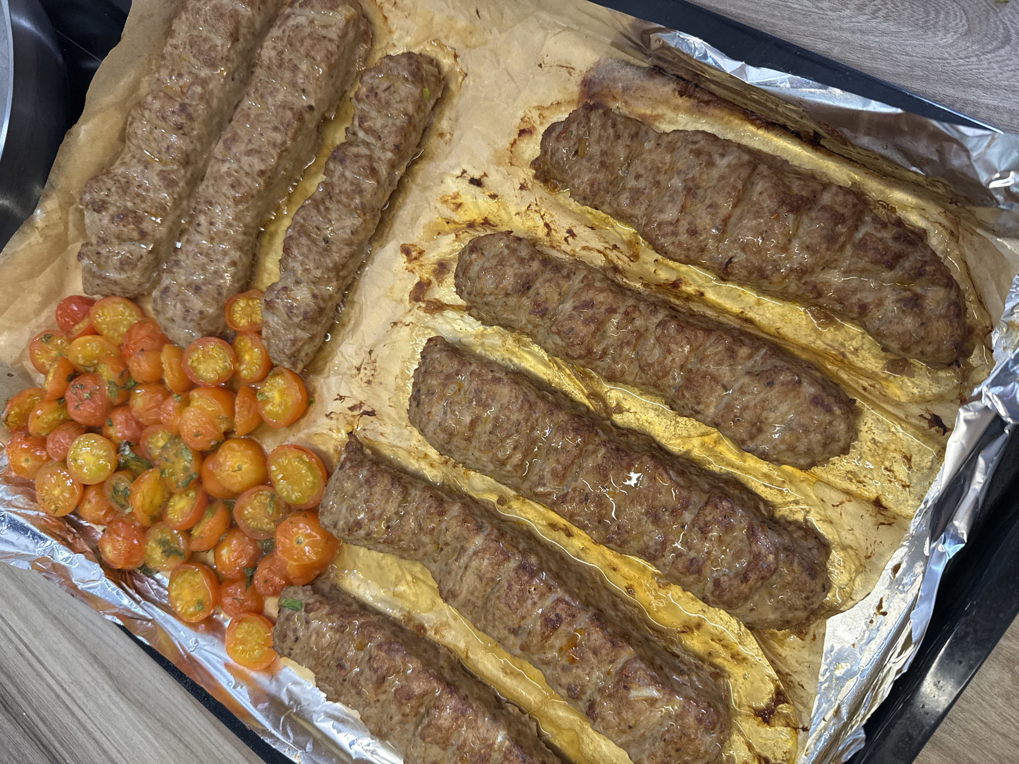 A top-down, close-up shot shows a baking sheet lined with parchment paper on a dark-colored baking pan, likely in a kitchen setting in Krasnoyarsk, Russia. The baking sheet contains several cooked, elongated meat skewers (likely lula kebab or similar) and halved cherry tomatoes. The skewers are arranged in parallel rows, some with visible oil or cooking juices. The cherry tomatoes appear to be seasoned. The scene is well-lit, emphasizing the texture and color of the food items.