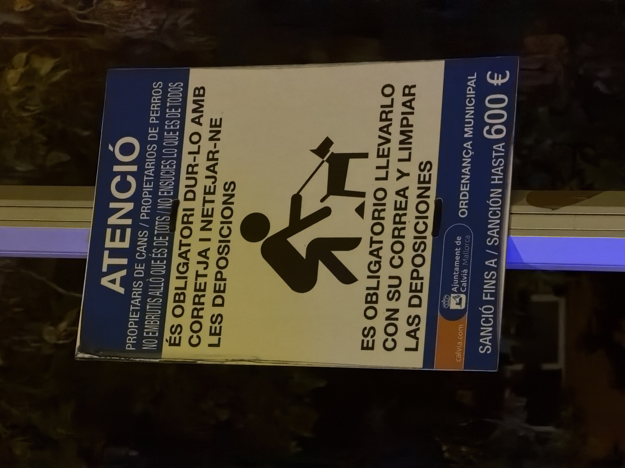 The image shows a sign with text in Catalan and Spanish, warning about the responsibility of dog owners. The sign is located on a pole, with a dark, out-of-focus background that suggests it is nighttime. The sign is yellow and blue, with a black pictogram of a person with a dog on a leash. The text in Catalan reads "ATENCIÓ PROPIETARIS DE CANS / PROPIETARIOS DE PERROS NO EMBRUTIS ALLÒ QUE ÉS DE TOTS / NO ENSUCIES LO QUE ES DE TODOS ÉS OBLIGATORI DUR-LO AMB CORRETJA I NETEJAR-NE LES DEPOSICIONS". This translates to "ATTENTION DOG OWNERS / OWNERS OF DOGS DO NOT FOUL WHAT BELONGS TO EVERYONE IT IS MANDATORY TO WALK IT ON A LEASH AND CLEAN UP ITS DEPOSITS". Below this, in Spanish, it reads "ES OBLIGATORIO LLEVARLO CON SU CORREA Y LIMPIAR LAS DEPOSICIONES". This translates to "IT IS MANDATORY TO WALK IT ON ITS LEASH AND CLEAN UP THE DEPOSITS". At the bottom of the sign, there is text in a blue bar which reads "calvia.com Ajuntament de Calvià Mallorca ORDENANÇA MUNICIPAL SANCIÓ FINS A / SANCIÓN HASTA 600 €". This indicates the sign is from the municipality of Calvià, Mallorca, and that the fine for non-compliance is up to 600 Euros. The overall scene depicts a public notice in a Spanish municipality, likely in a park or on a street, aimed at ensuring public cleanliness by enforcing responsible dog ownership.